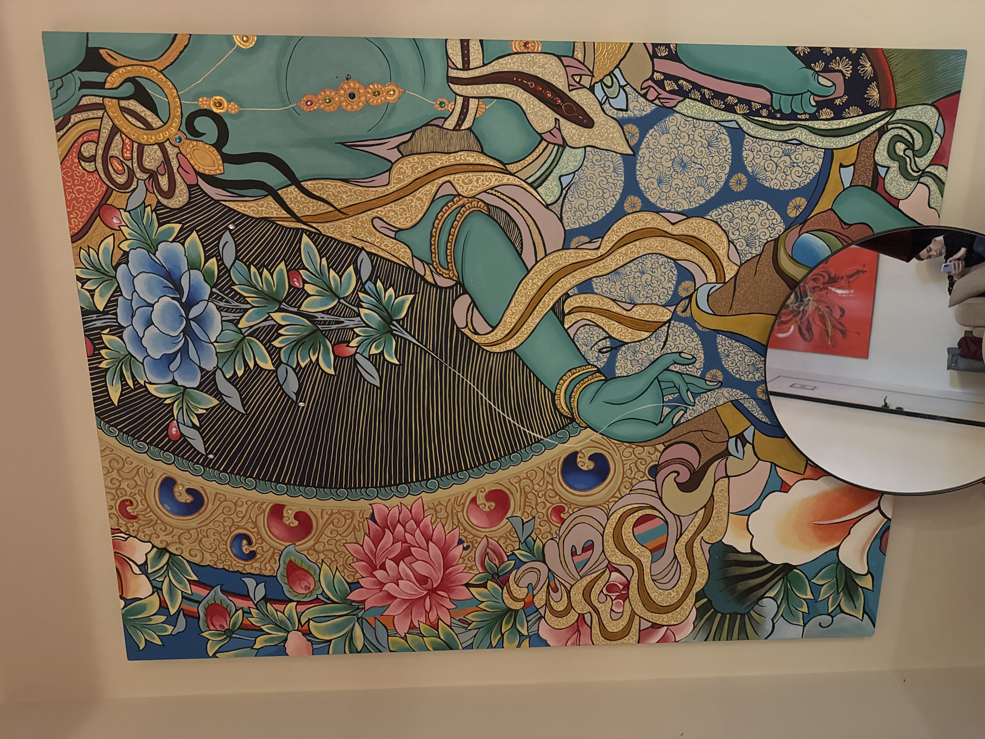 The image shows a colorful mural on a wall. The mural is dominated by an aqua-skinned figure with gold embellishments, surrounded by stylized flowers and patterns. There is a blue flower in the lower left corner, a pink flower in the lower center, and an orange and white flower in the lower right corner. A round mirror in the lower right corner reflects an adjacent room with a red artwork and a person holding a phone. The location of the mural is in Krasnoyarsk, Russia.