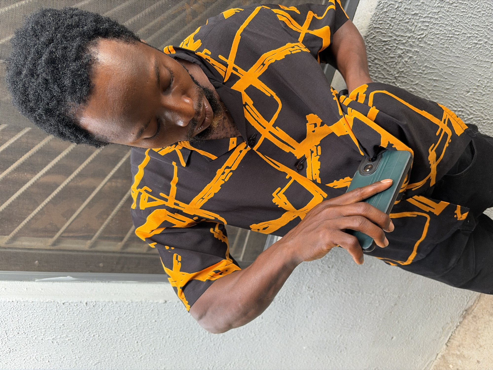 A high-angle medium shot depicts a man holding a dark teal smartphone. He is dark-skinned, with short, dark, tightly curled hair. His eyes are closed, and he is looking down at the phone in his right hand. He wears a short-sleeved button-up shirt with a black background and abstract orange geometric patterns. He also wears black pants.
The background includes a white, textured surface which appears to be a wall. Above the wall is a metal security screen. The location is likely Jos, Nigeria.