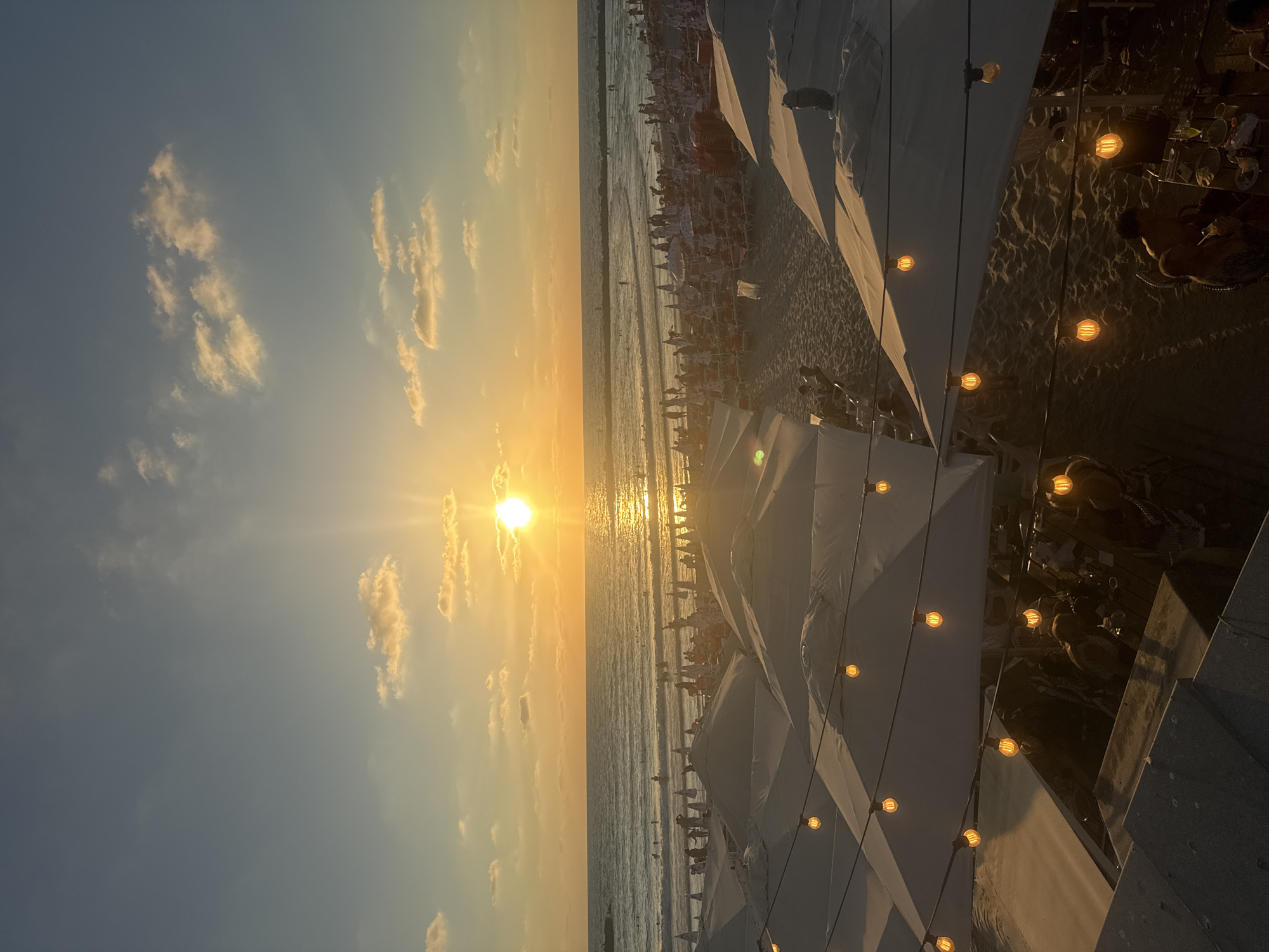 This is a wide-angle, eye-level shot of a crowded beach scene in Tel Aviv, Israel, during sunset. The sun, a bright orb, is setting over the Mediterranean Sea, casting a warm, golden glow across the sky and water. The sky is a gradient of blue and orange, with scattered clouds tinged with yellow from the setting sun.
In the foreground, a series of large, white beach umbrellas are set up on the sand, some partially obscuring the view. String lights with visible Edison bulbs are suspended above these umbrellas, adding a festive ambiance. People are seen under and around these umbrellas, some seated at tables, suggesting a beachside dining or lounge area.
Beyond the umbrellas, the sandy beach is packed with people. Many are lounging on beach chairs under smaller, red-and-white striped umbrellas. Other figures are walking along the shore or standing in small groups.
The sea is a deep blue, with gentle waves rolling towards the shore. Several individuals are visible in the water, swimming or wading. A breakwater or pier-like structure is visible in the distance, breaking up the horizon line. The overall impression is a lively and popular public beach enjoying the end of a warm day.