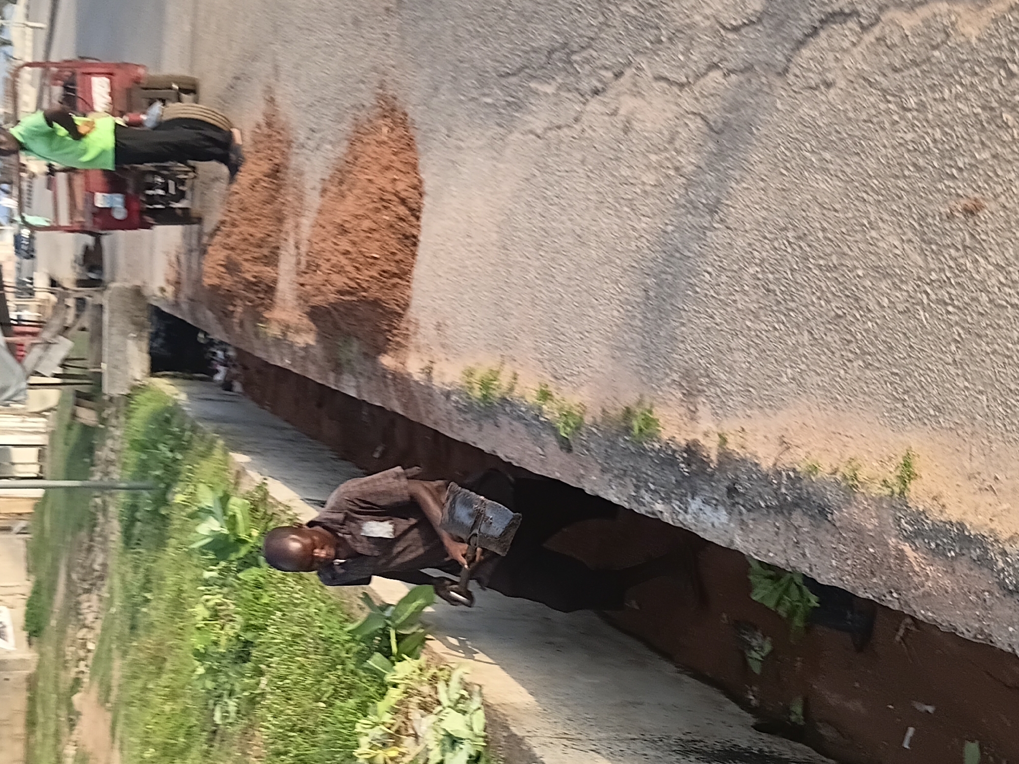 In Jalingo, Nigeria, a man is working in a concrete drainage ditch next to a paved road. He is wearing a brown checkered shirt and dark pants and is using a shovel and a dustpan to clear debris from the ditch. The ditch contains water and plant matter. On the road next to the ditch, there are piles of reddish-brown soil. A red three-wheeled vehicle is parked on the side of the road, and another person in a bright green shirt is standing next to it. Green vegetation is visible on the left side of the image, adjacent to the ditch. The sun is casting shadows, suggesting it is daytime. The overall scene depicts manual labor and infrastructure maintenance.