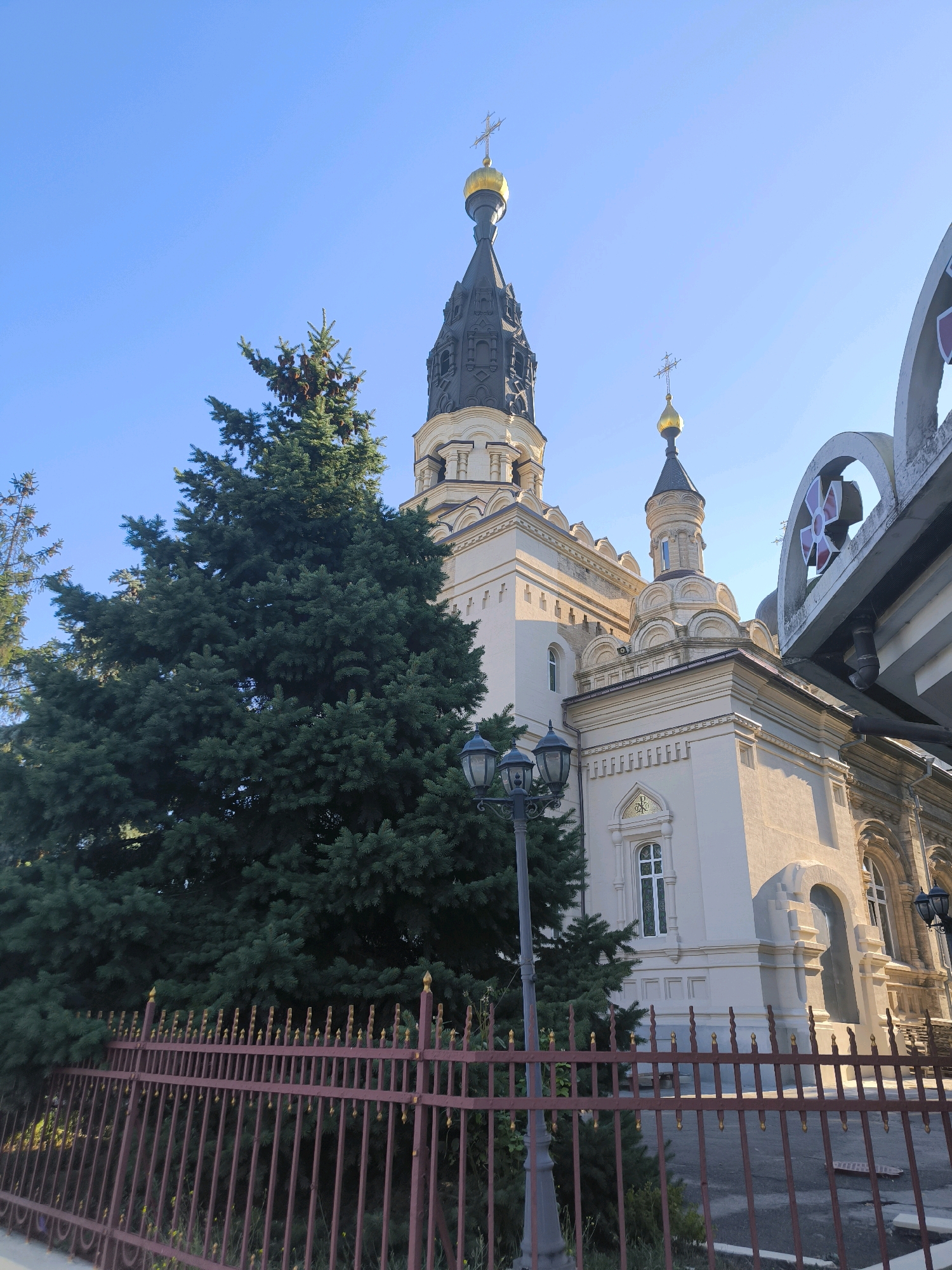 The image depicts the Church of the Assumption of the Blessed Virgin Mary, located in the city of Mykolaiv, Ukraine. The church is a prominent religious building with a central dark spire topped with a golden dome and cross. Adjacent to it is another smaller dome, also golden, with a cross. The church's facade is light-colored, featuring arched windows and decorative architectural elements.
In the foreground, a dark red metal fence with pointed finials runs horizontally across the lower part of the image. Behind the fence, a large, lush green coniferous tree dominates the left side of the frame, its branches full and dense. A black lamppost with multiple lights stands next to the tree.
The sky is a clear, bright blue, indicating a sunny day. The lighting suggests it is late afternoon, with the sun casting shadows and highlighting the textures of the building and tree. The overall scene is tranquil and well-maintained, showcasing a religious landmark within an urban or suburban environment. There are no people visible in the image.