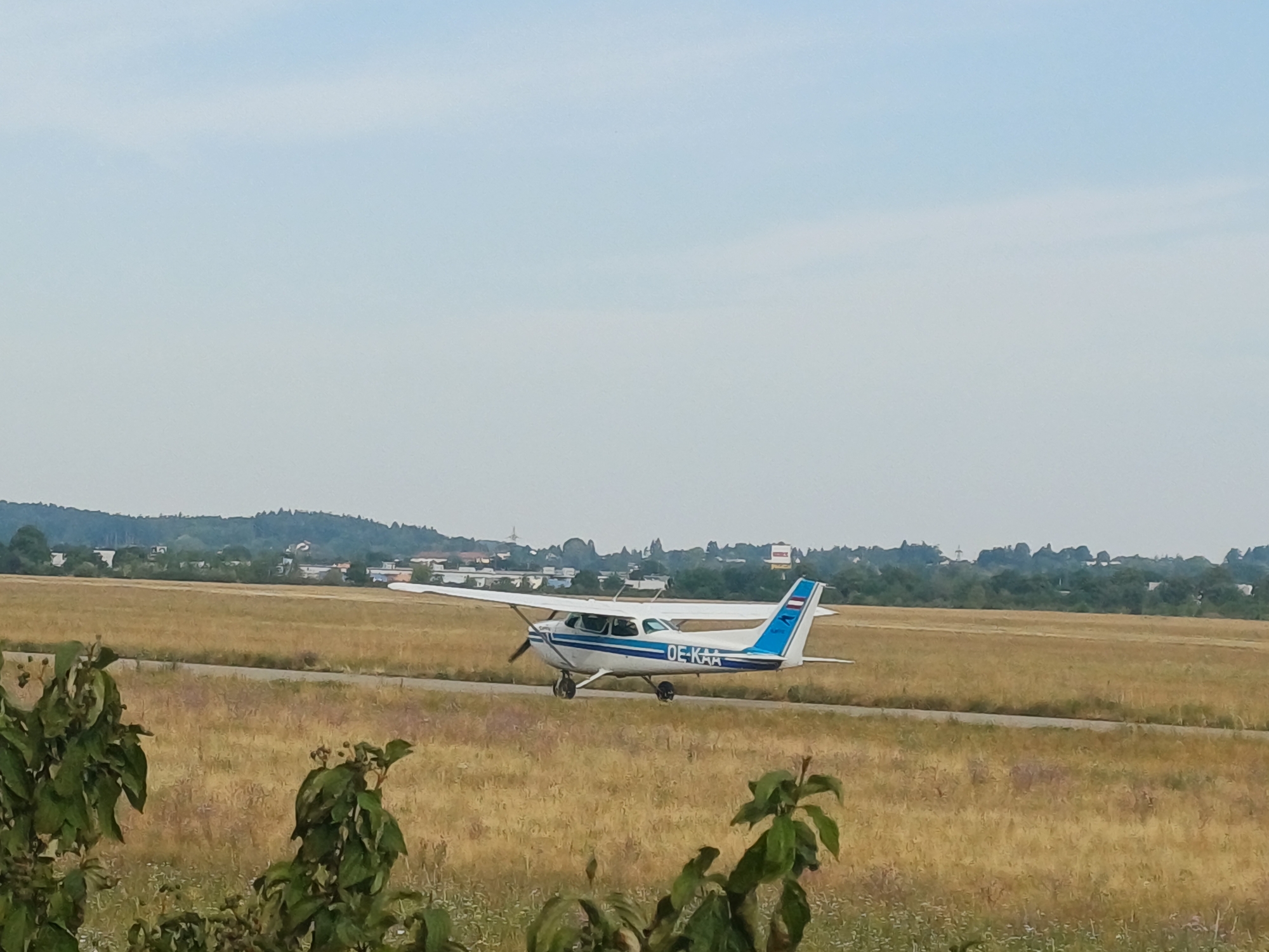 This is a medium shot of a small, single-engine airplane on a tarmac at an airfield. The airplane is white with blue stripes and the registration "OE-KAA" is visible on the fuselage. It is positioned on a paved runway that cuts through a dry, golden-brown field. In the background, a line of trees and buildings can be seen, suggesting a rural or suburban area. The sky is a pale blue with some wispy clouds. The foreground shows blurred green foliage. This scene is likely at the Wels Airfield (Flughafen Wels) in Wels, Austria, a general aviation airport. The plane appears to be stationary or taxiing.