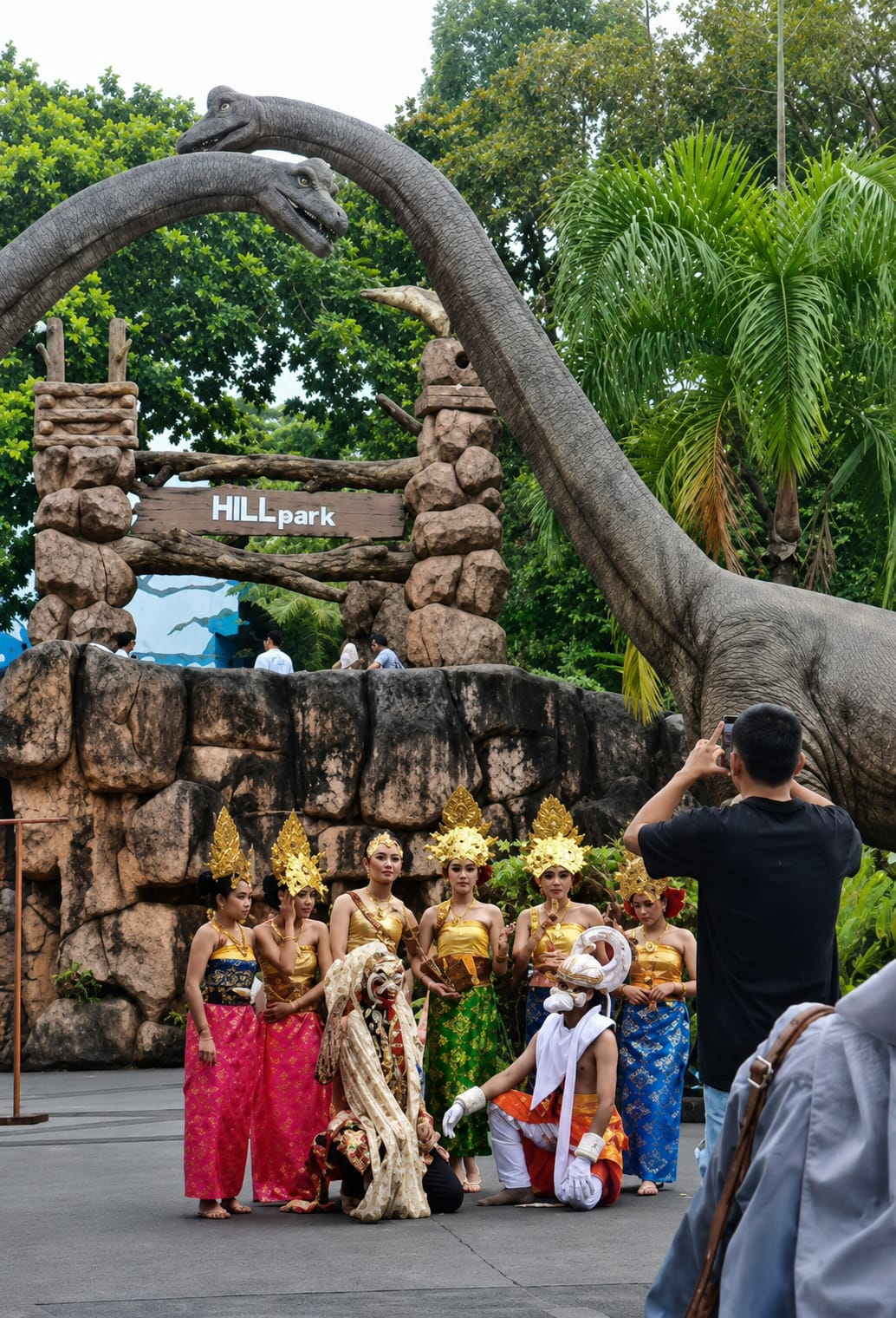A group of performers in traditional costumes, including women in ornate gold headdresses and colorful sarongs, and two masked figures, pose for a photograph in front of a stone facade and a large dinosaur statue at Hill Park in Bandar Baru, Indonesia. A man in a black t-shirt is seen taking a photo of the performers with his phone. The background features lush green trees and a cloudy sky.