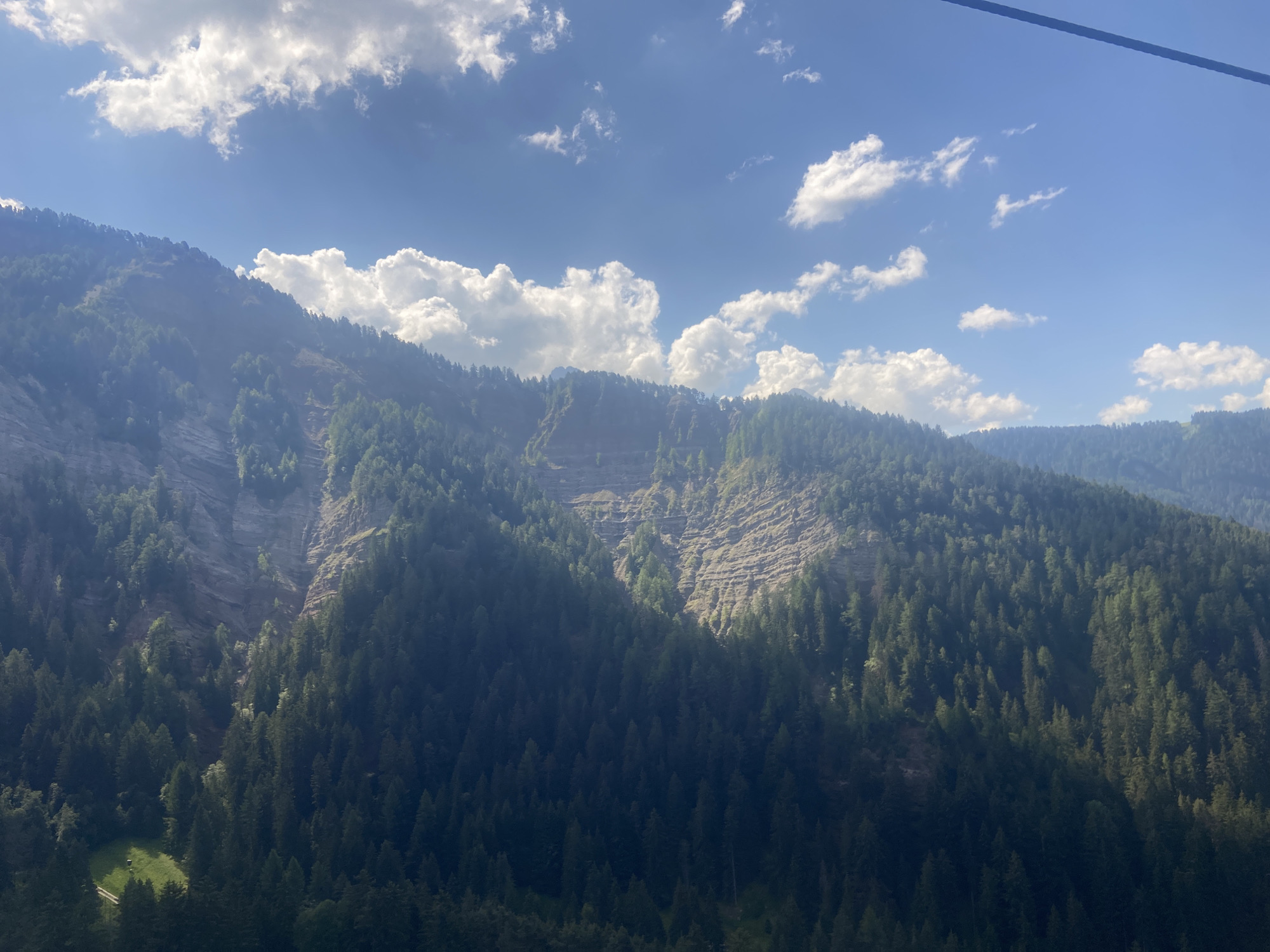 The image depicts a panoramic view of a mountainous landscape, likely captured from a high vantage point such as a cable car or gondola, given the presence of a cable running across the top of the frame. The scene is dominated by lush green evergreen forests covering the steep slopes of mountains. A prominent rock face, characterized by terraced layers and scree, is visible on the left side of the frame, partially illuminated by sunlight. The sky is a vibrant blue, dotted with scattered white cumulus clouds. In the lower left portion of the image, a small clearing with a patch of bright green grass and a hint of a path can be seen within the dense forest. The overall impression is one of a serene and majestic natural environment. This scene is characteristic of the Val Gardena region in South Tyrol, Italy, where mountain resorts like Ortisei (Urtijëi - St. Ulrich) are situated.