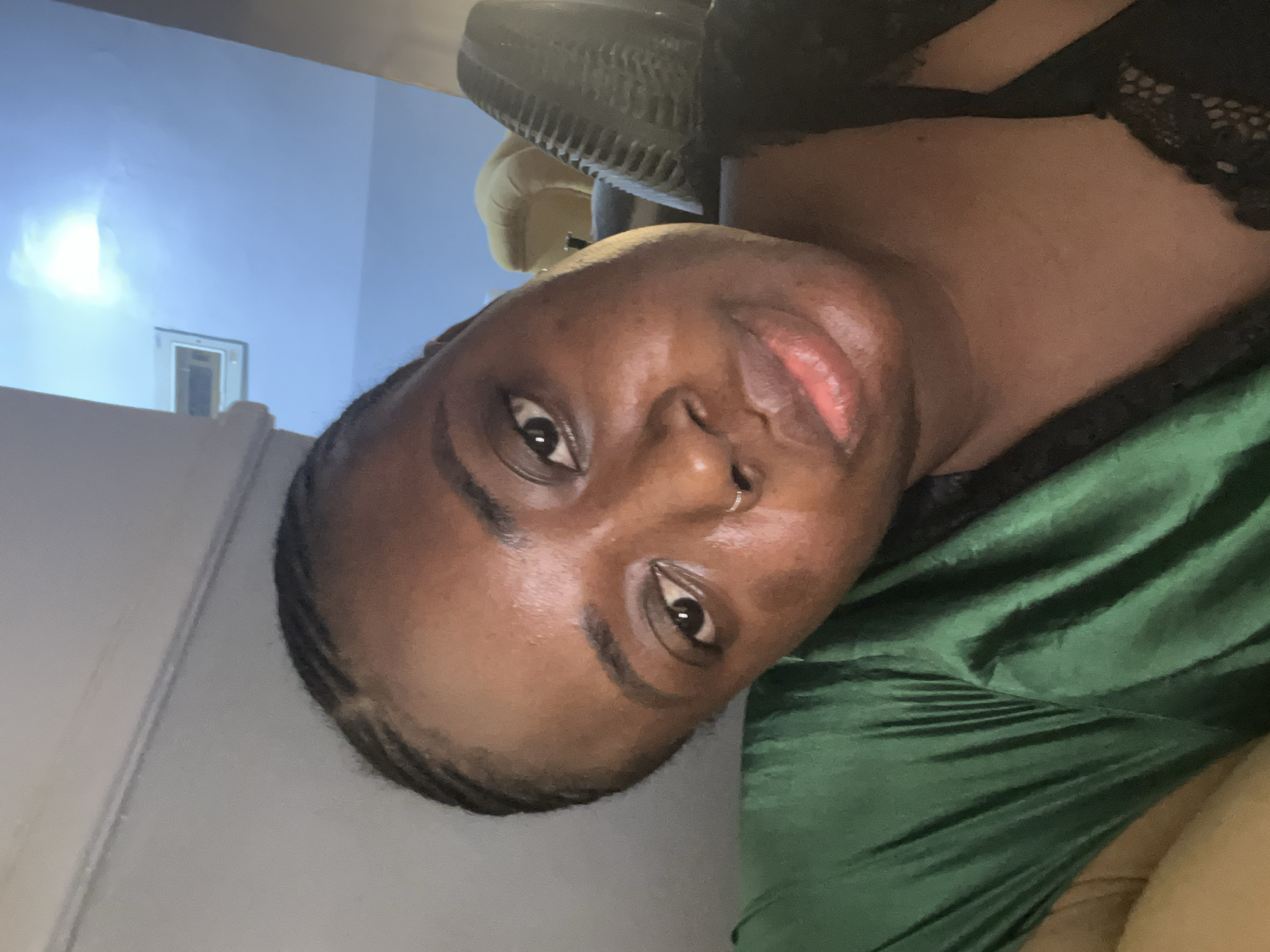 A close-up selfie shows a brown-skinned individual with dark hair pulled back in braids. They are wearing a green satin top and a black lace-trimmed garment beneath. A black, textured headrest of a chair is visible behind the individual. The background shows a white wall with an indistinct white rectangle. The location appears to be indoors, with natural light coming from a window. There is no indication of the city of Kubwa, Nigeria, within the frame of the photograph.