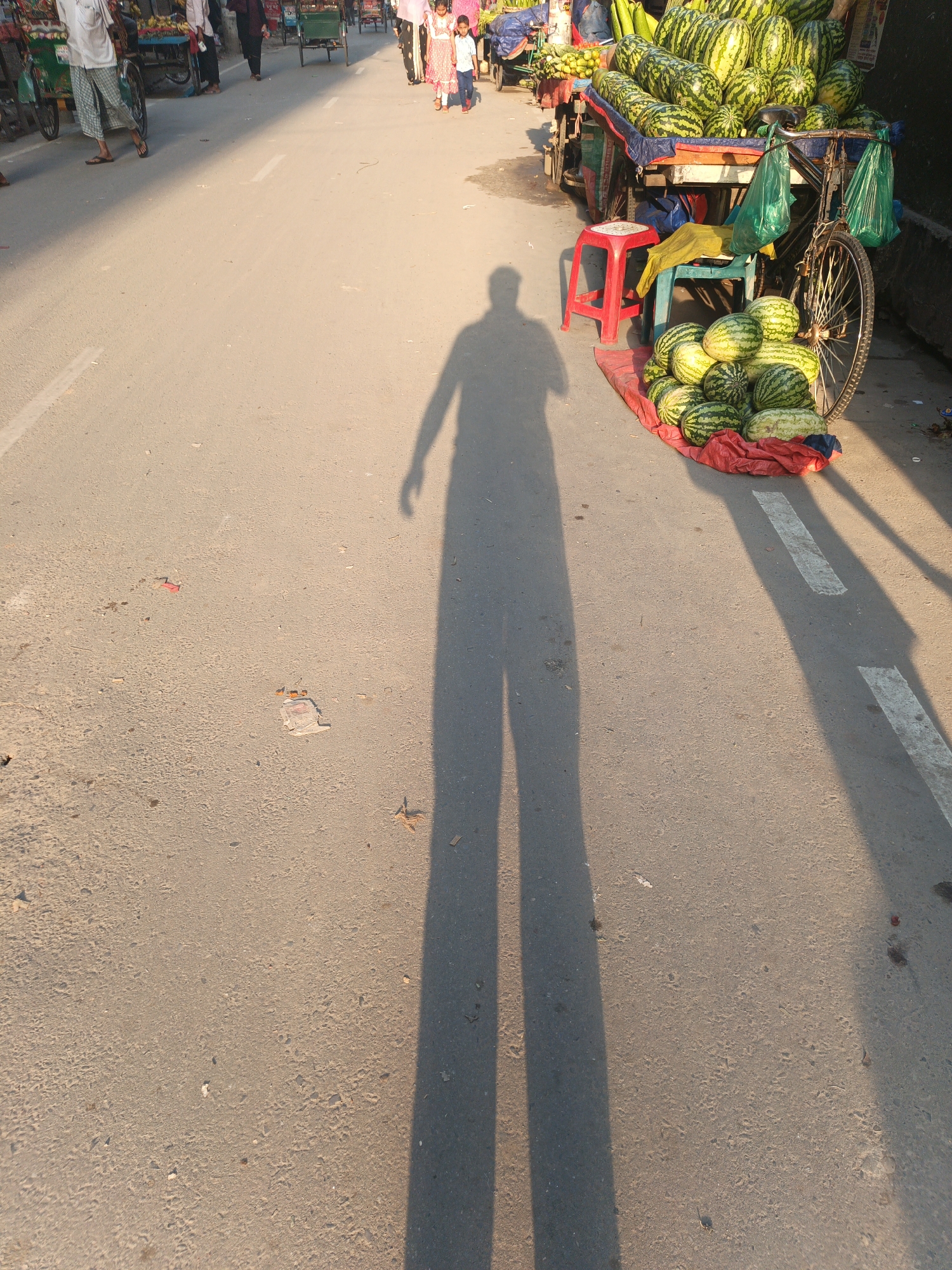 The image depicts a street scene in Dhaka, Bangladesh, characterized by intense sunlight casting a long shadow of a person onto the pavement. A roadside stall is visible, laden with watermelons, some displayed on a cart and others arranged on the ground. People are walking in the background. The street surface is worn, with minor debris scattered around. The stall area is cluttered with objects including stools.