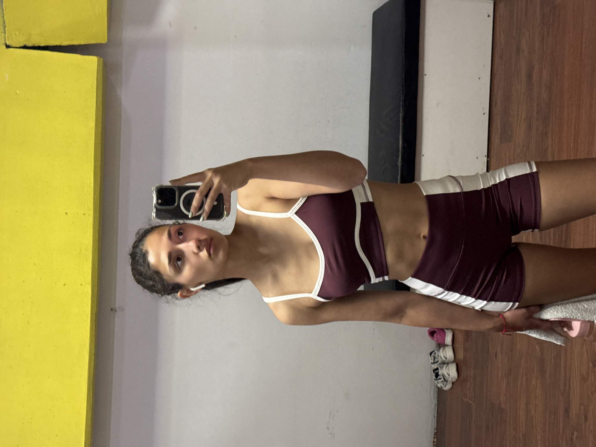 A young woman is captured in a mirror selfie, holding a smartphone with a clear case in her right hand. She looks directly at her reflection, positioned slightly to the left of the frame. Her dark hair is pulled back, and she wears white earbuds. She is dressed in maroon athletic wear, consisting of a sports bra and matching shorts, both featuring white trim and stripes. Her midriff is visible, and she wears a small red wristband on her left wrist.
The indoor setting appears to be a gym or a workout space. The woman stands on a wooden floor. In the background, bright yellow, rectangular block-like structures are visible on the left side, contrasting with a light grey wall that fills the central and upper right portions of the frame. A vertical black object, possibly a piece of equipment or a structural column, is visible behind her right arm. On the wooden floor to her right, a pair of light-colored sneakers is visible next to a white towel that she is gently holding with her left hand. Her expression is neutral.
The scene conveys a moment of self-reflection, likely before or after a workout. There is no visible text in the image. The location context is Šušanj, Montenegro.