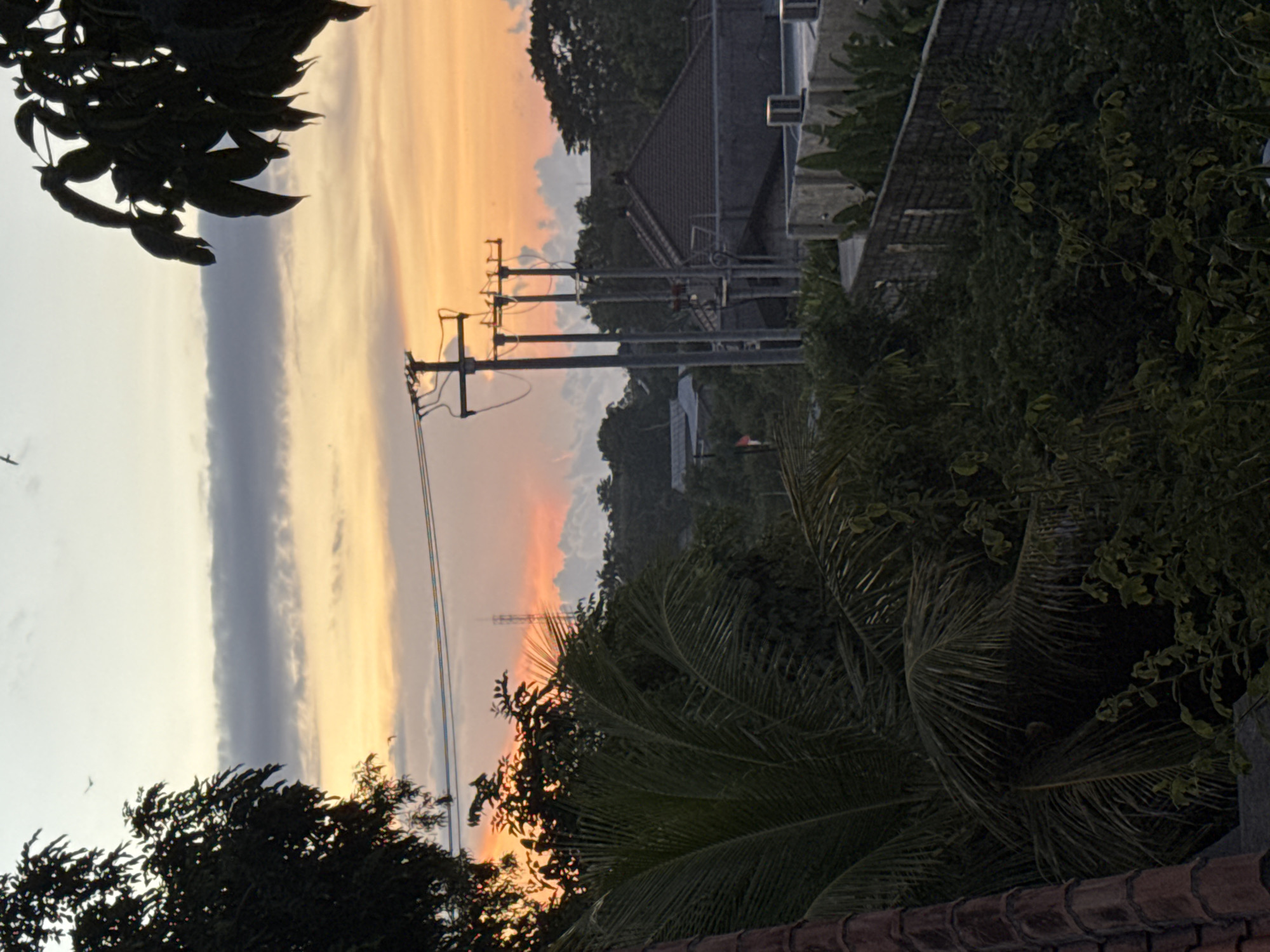 This media file captures an outdoor landscape scene at either sunrise or sunset in Kuta Selatan, Indonesia. The sky dominates the upper portion of the image, displaying a striking gradient of colors. Towards the right side, warm hues of orange and yellow blend into a lighter, paler sky that transitions to light grey and pale blue on the left. A prominent, long, dark grey cloud band stretches horizontally across the mid-section of the sky.
In the midground, a series of utility poles with multiple cross arms and wires are sharply silhouetted against the vibrant sky. Behind these poles, several residential buildings with dark, pitched roofs are partially visible, nestled among dense trees.
The foreground features lush, dark green foliage, including discernible palm fronds, particularly in the bottom right corner. A dark, tiled roof edge runs along the very bottom of the frame. In the far left, silhouetted tree branches with leaves extend into the view from the top corner.
In the distant background, rolling hills covered in dense, dark vegetation are visible, fading into the hazy horizon. A faint silhouette of a tall tower or pylon can also be seen against the colorful sky in the distance. The overall mood conveyed by the warm sky colors and tranquil setting is peaceful. There are no visible people or discernible text.