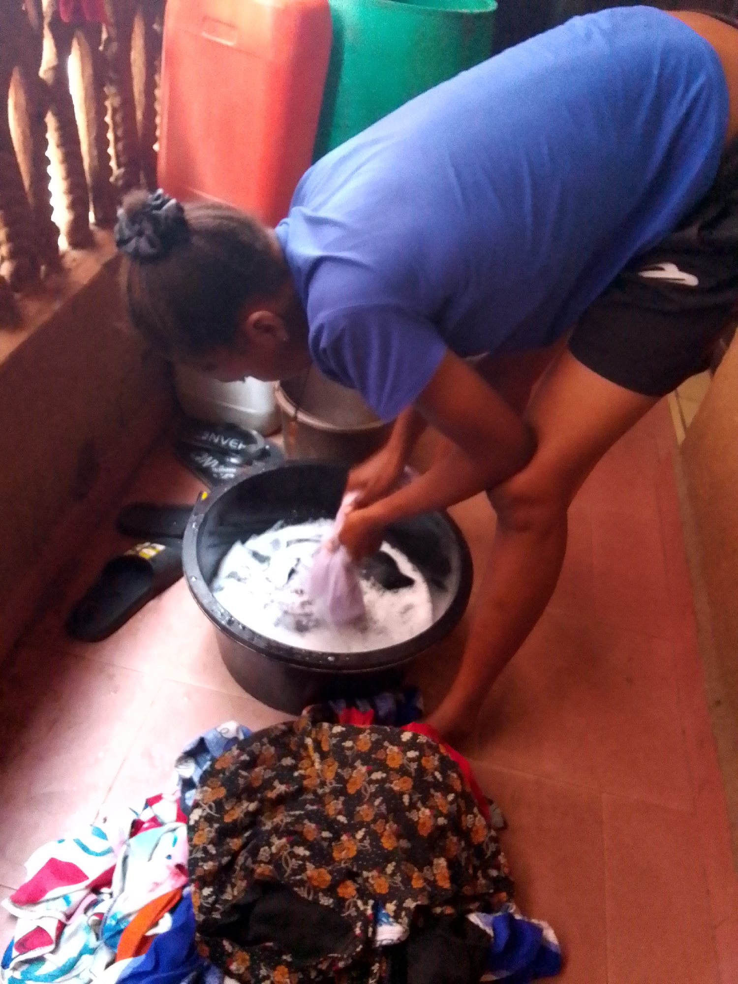 A young woman is hand-washing clothes in a black basin filled with soapy water. She is wearing a blue t-shirt and black shorts, and her hair is tied back in a bun. Her posture suggests she is bent over the basin, actively scrubbing or rinsing the clothes. A pile of clean, folded clothes sits on the ground near the basin. The setting appears to be an indoor or semi-indoor space with a tiled floor. In the background, there are several large, colorful containers and a fence. This image was taken in the city of Unknown, Unknown.
