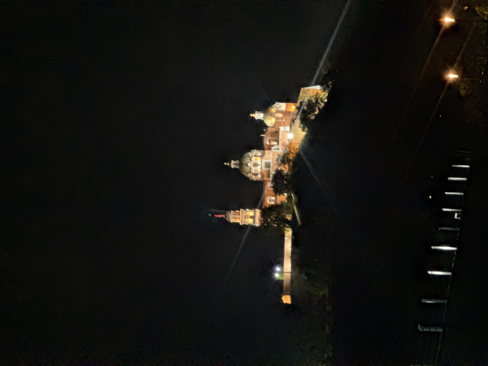 This is a nighttime shot of Santuario de la Virgen de los Remedios in San Andrés Cholula, Mexico. The illuminated church, with its domes and bell tower, is the central focus. A bridge or pathway leading up to the church is also lit up. The surrounding environment is dark, and the lighting creates strong contrasts. To the right, there are streetlights and structures along the water's edge.