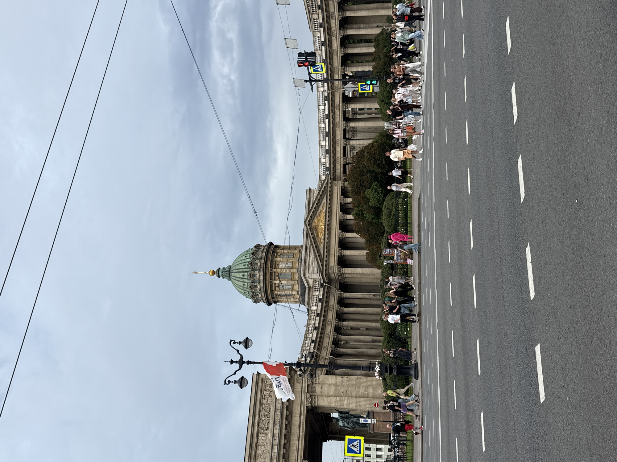 The image depicts a wide street in Saint Petersburg, Russia, with Kazan Cathedral prominently featured in the background. The cathedral is a large, neoclassical building with a distinctive green dome and a semi-circular colonnade. Several people are crossing the street at a pedestrian crossing, with traffic lights visible. A lamppost with two lamps and a flag is located on the left side of the street. The sky is overcast. The scene is a busy urban environment.