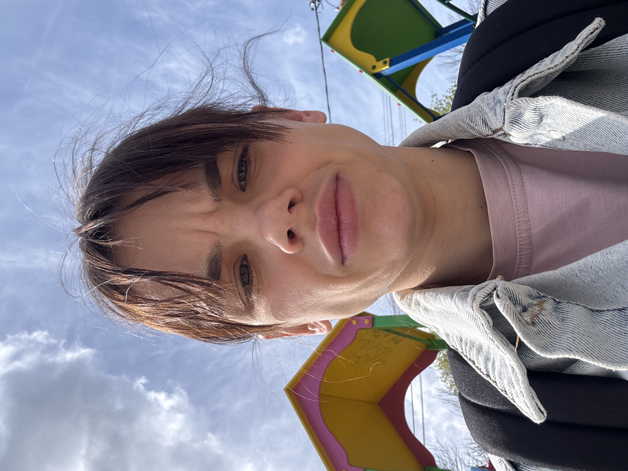 A close-up, tilted selfie features a person with short brown hair and fair skin, wearing a pink t-shirt and a light-wash denim jacket over what appears to be a black backpack. The background is a bright sky with scattered clouds and colorful playground equipment, suggesting a park setting. The person's expression is a slight frown or grimace. Without additional information, the specific location in Rostov-on-Don cannot be determined.