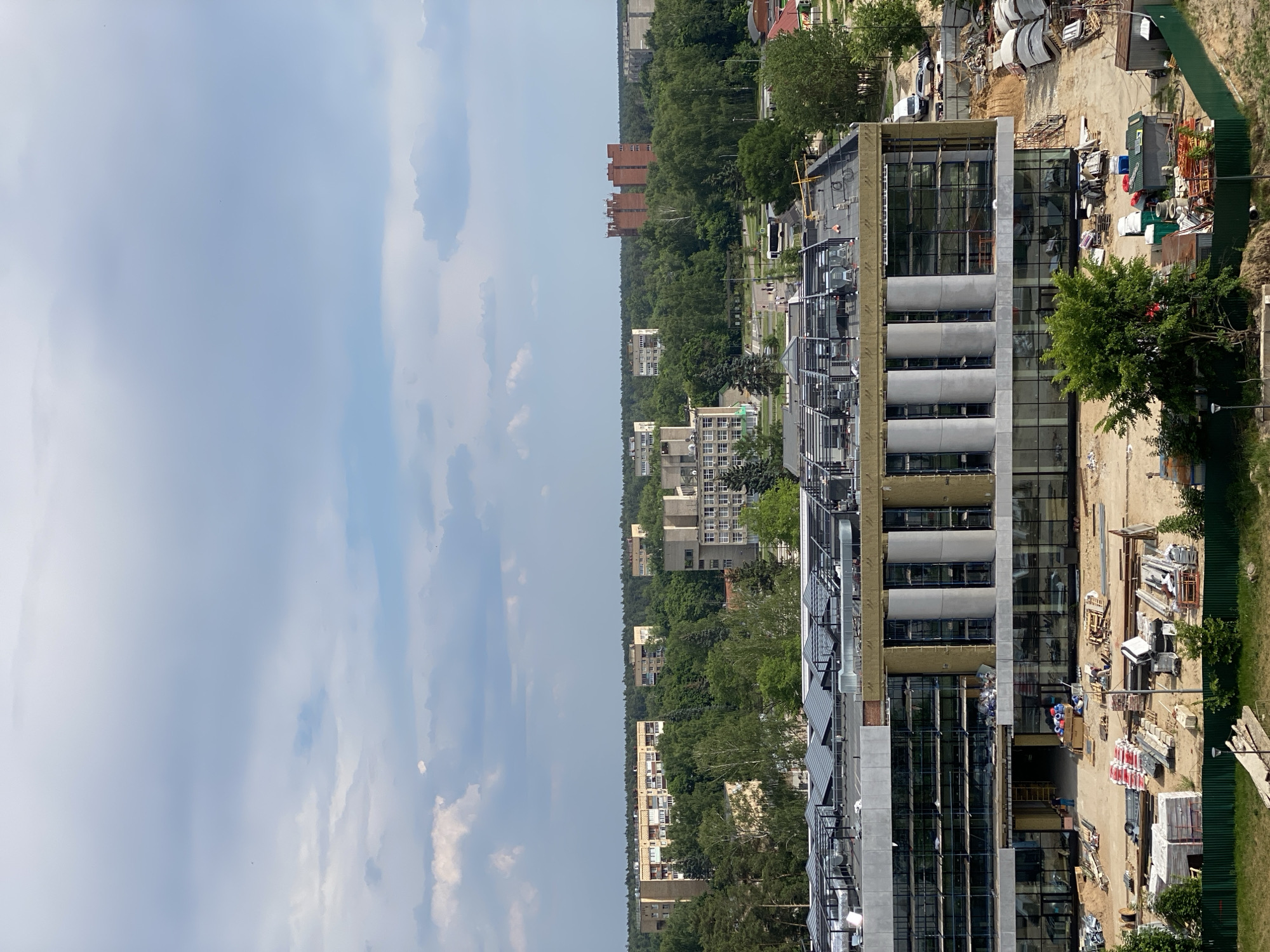This is a vertical, medium-distance, aerial shot of a city under construction. The foreground features a partially constructed building with large glass windows and exposed insulation. Construction materials, including stacks of lumber, pipes, and pallets of supplies, are scattered around the site. Several portable toilets and small sheds are also visible. A green metal fence encloses the construction area. In the middle ground, a modern building with a flat roof is under construction, with visible scaffolding and exposed structural elements. Beyond this, a dense forest stretches across the horizon, dotted with residential buildings of varying architectural styles, including apartment blocks and some older brick structures. A red brick building with multiple stories stands out among the trees in the distance. The sky is partly cloudy with patches of blue. The overall impression is of an urban area undergoing development amidst a natural landscape.