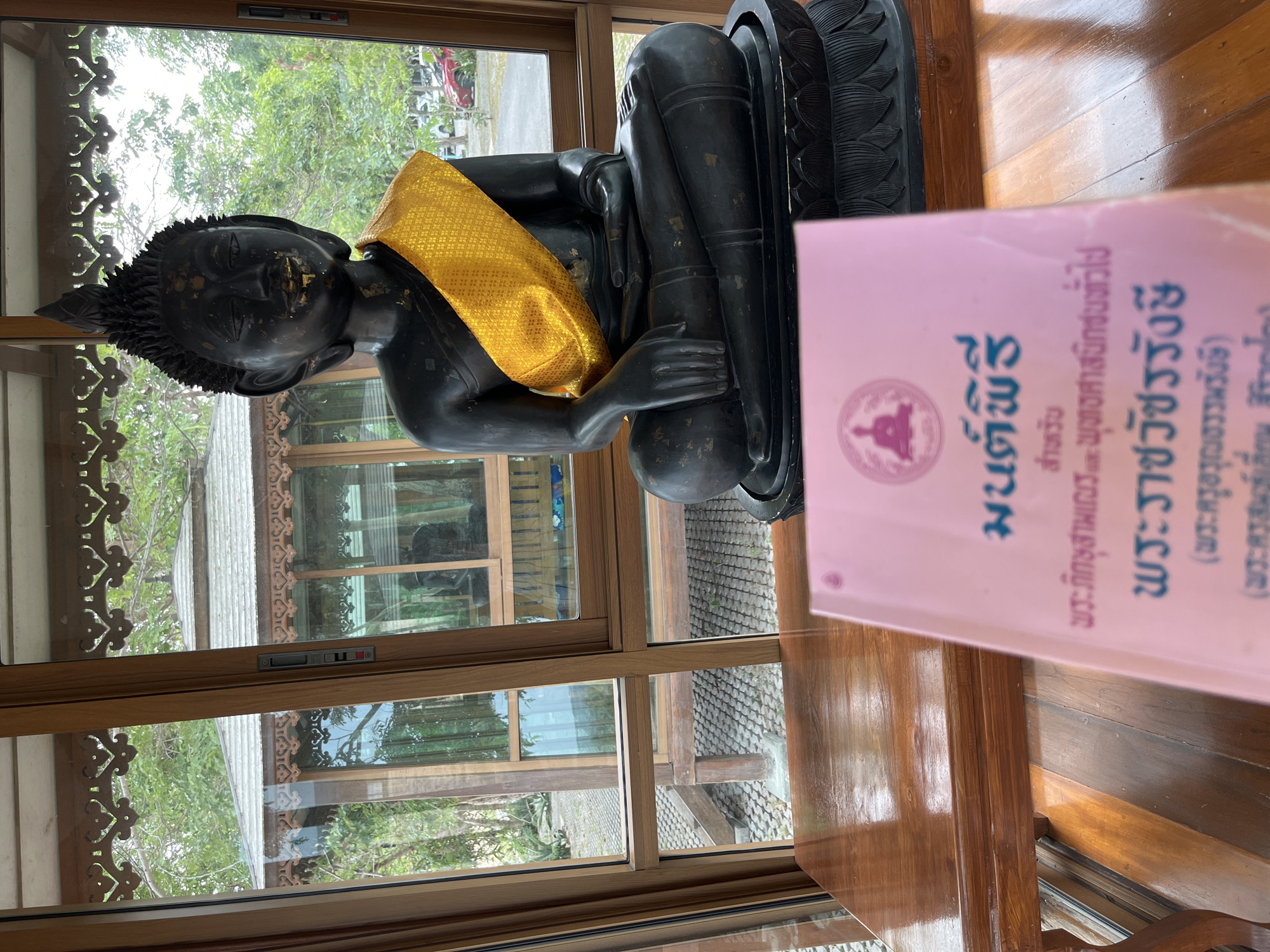 A statue of a seated figure with a gold sash, likely Buddha, sits on a wooden surface near a window. A pink book with Thai script partially obscures the view of the statue. The setting appears to be inside a building with views of trees and other structures visible through the window. The location is unspecified.