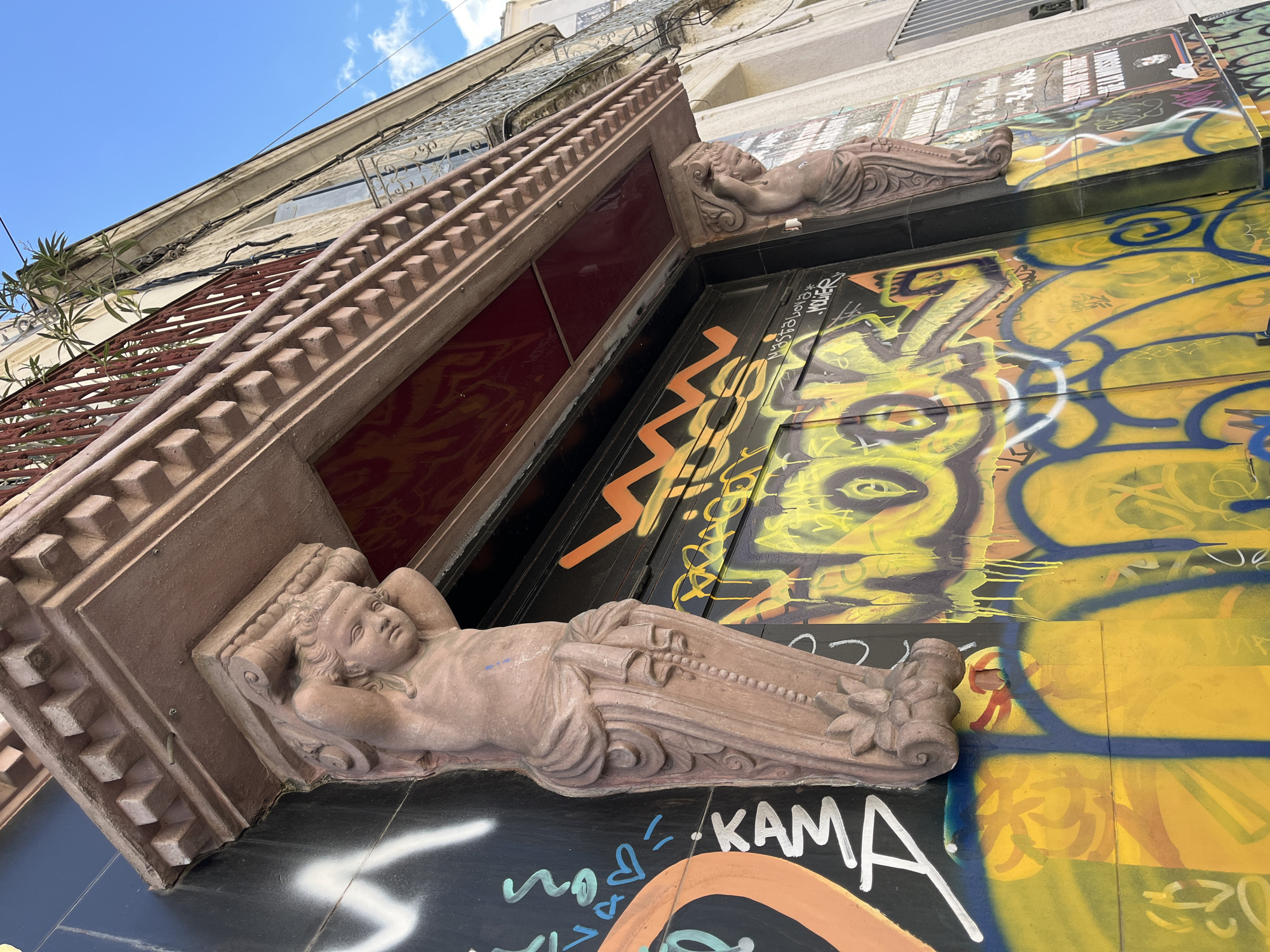The image shows the facade of a building in Montpellier, France, featuring architectural details and graffiti. The entrance is flanked by two sandstone sculptures of reclining figures, possibly putti or angels. The facade above the entrance is covered in layers of graffiti, mainly in yellow, orange, blue, and black. The building itself appears to be old, with a balcony with wrought-iron railings visible in the upper left of the frame. The sky is blue with some scattered clouds.