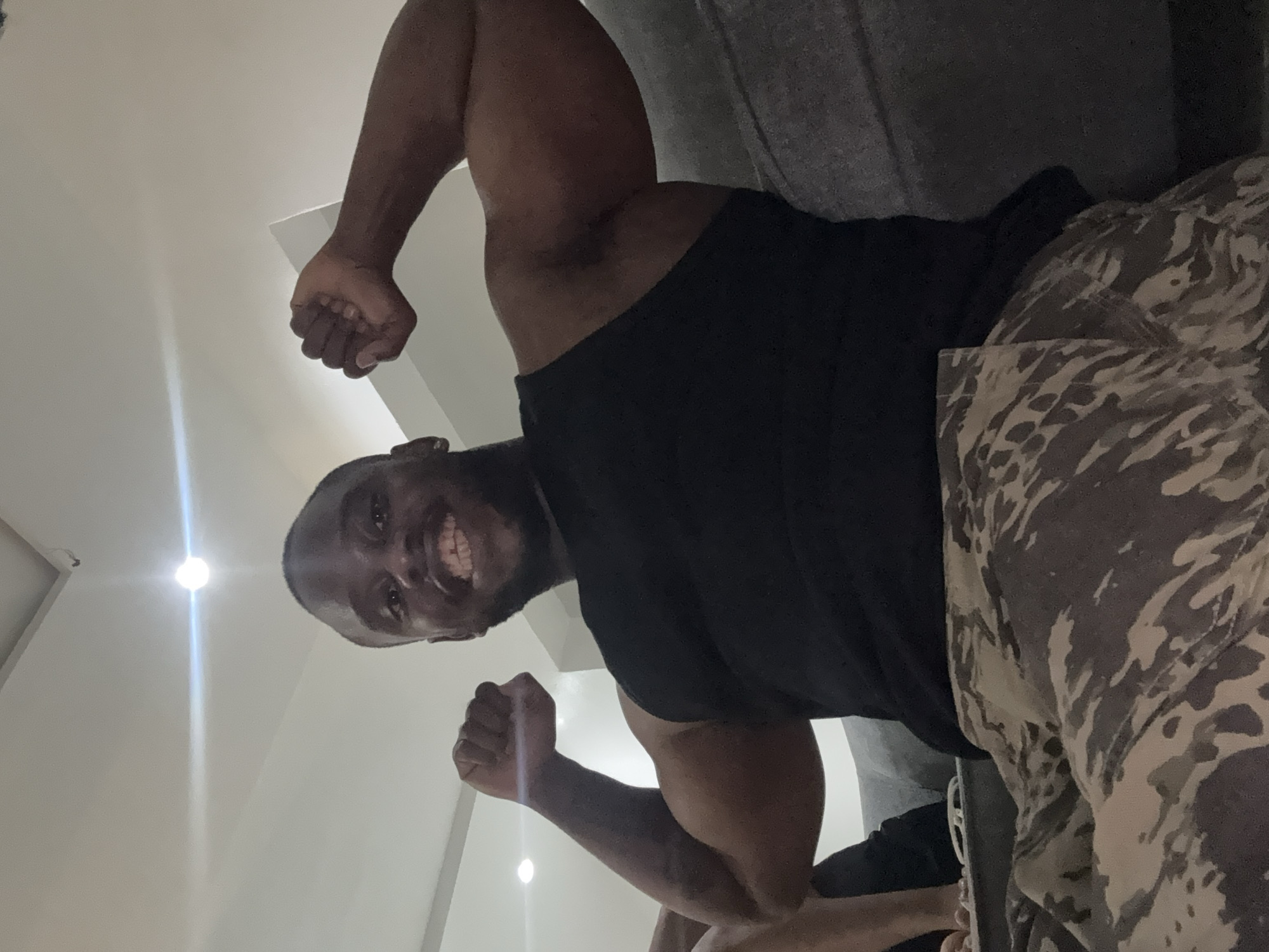 The image features a dark-skinned man captured from a low-angle perspective, looking down towards the camera with a wide, joyful smile that reveals his teeth. He is actively flexing both of his bicep muscles, with clenched fists, conveying a sense of strength and confidence. He is dressed in a dark, likely black, short-sleeved t-shirt and camouflage-patterned shorts or trousers, featuring shades of grey, beige, and brown.
The scene is set indoors, with a plain, light-colored background that appears to be a wall or ceiling. Two bright, artificial light sources, possibly recessed ceiling lights, are visible in the background, creating noticeable lens flares and light streaks. A portion of a dark grey or black piece of furniture, potentially a couch, can be seen along the right edge of the frame. The overall mood conveyed by the man's expression and pose is one of happiness and robust vitality. No discernible text is visible within the image.