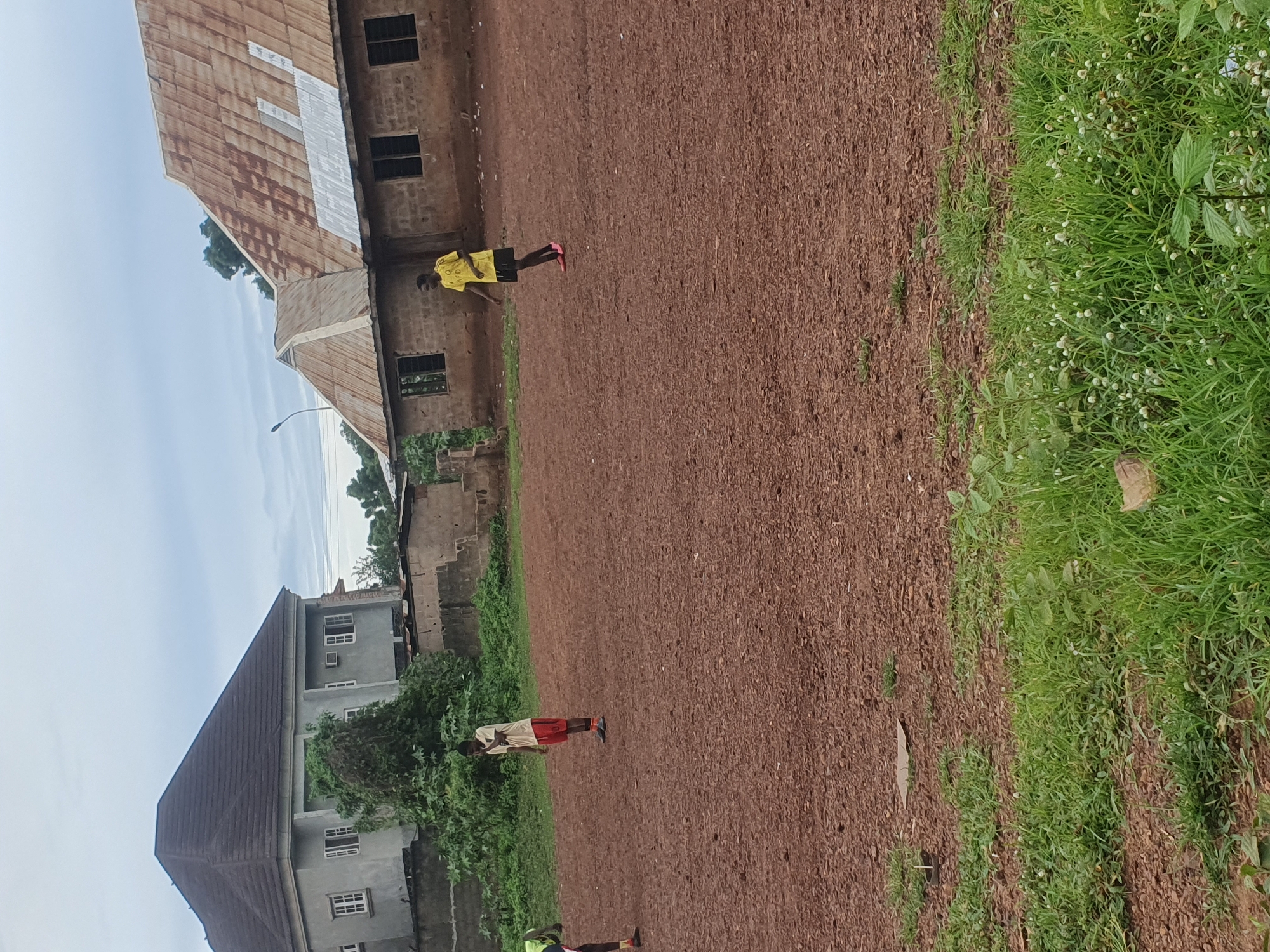 The image depicts an outdoor, semi-urban scene in Idah, Nigeria, under an overcast sky. The foreground and midground consist of an unpaved, reddish-brown dirt expanse, sparsely covered with small rocks and sporadic patches of green grass. In the lower-left foreground, a small, dark cylindrical object is visible in the grass.
Three individuals are present on the dirt field. On the far left, partially cropped, is an individual wearing a green or yellow top and dark shorts, with a dark shoe visible. In the center of the midground, a male wearing a light-colored t-shirt and orange-red shorts stands upright, facing right, with his hands near his face. To the right, another male, wearing a yellow t-shirt with a dark logo (partially reading "AFO") and black shorts, with bright pink shoes, walks from right to left.
In the background, two distinct structures are visible. On the left, a multi-story building with light grey walls and a dark, multi-pitched roof stands. It features multiple windows with white frames and grilles, and an external air conditioning unit on an upper wall. The base of this building is partially obscured by dense green foliage. On the right, a single-story building constructed with unpainted brick walls and a multi-pitched roof made of corrugated metal sheets, displaying both rusted brown and newer silver sections, is present. This structure also features multiple windows with dark frames and grilles. Additional green vegetation is visible between and behind the buildings, and a utility pole is faintly discernible on the horizon to the far right.