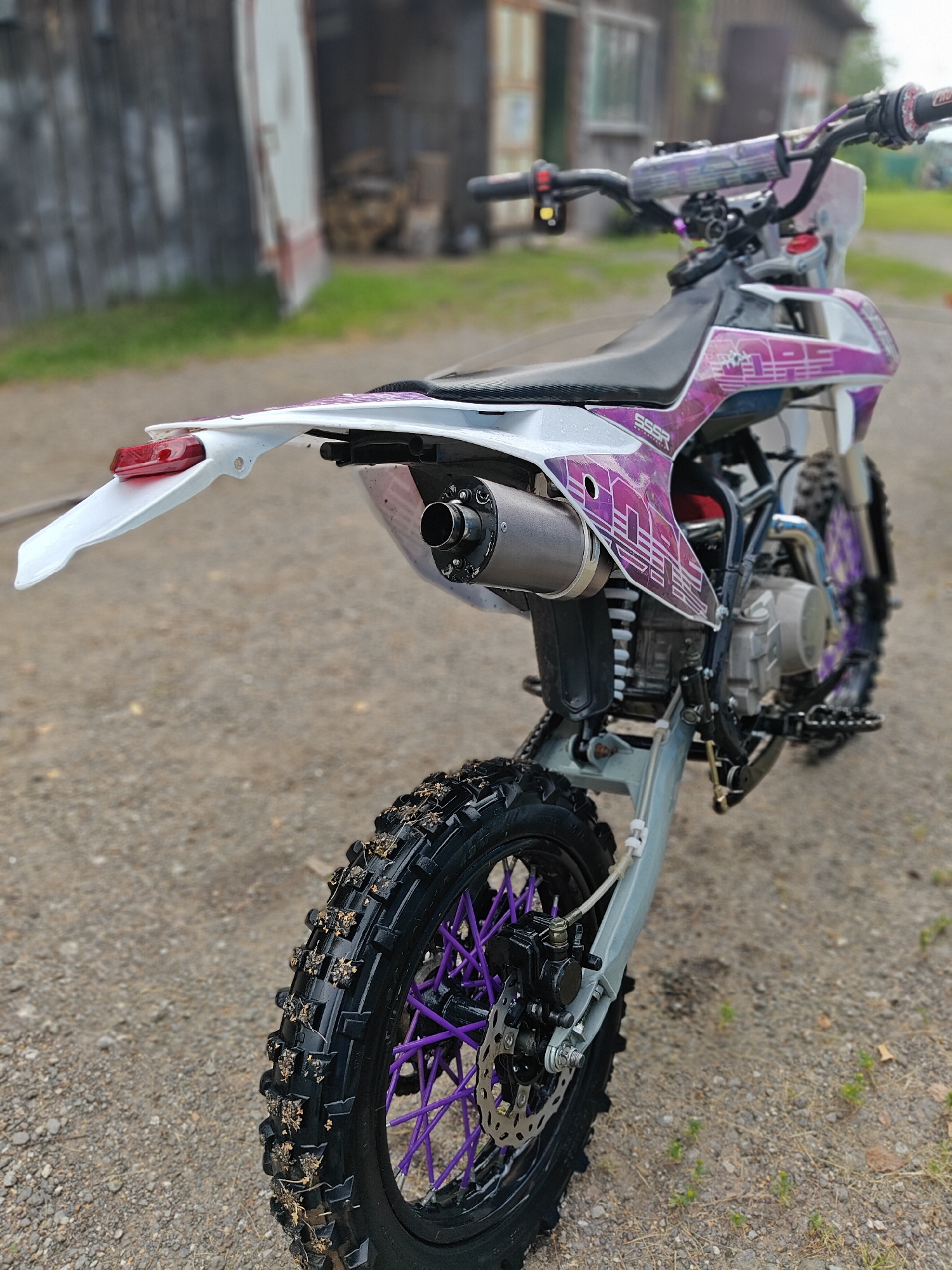 A dirt bike is parked on a gravel surface in front of a weathered wooden building. The motorcycle is predominantly white and purple with black tires and purple spokes. The rear tire is prominently featured, with mud and debris clinging to the tread. The building in the background appears to be a shed or barn, with stacked firewood visible. The scene is outdoors, likely in a rural setting, given the unpaved ground and rustic architecture. The lighting suggests it is daytime, possibly overcast due to the soft shadows. The overall impression is one of a functional, off-road vehicle at rest in a utilitarian environment.