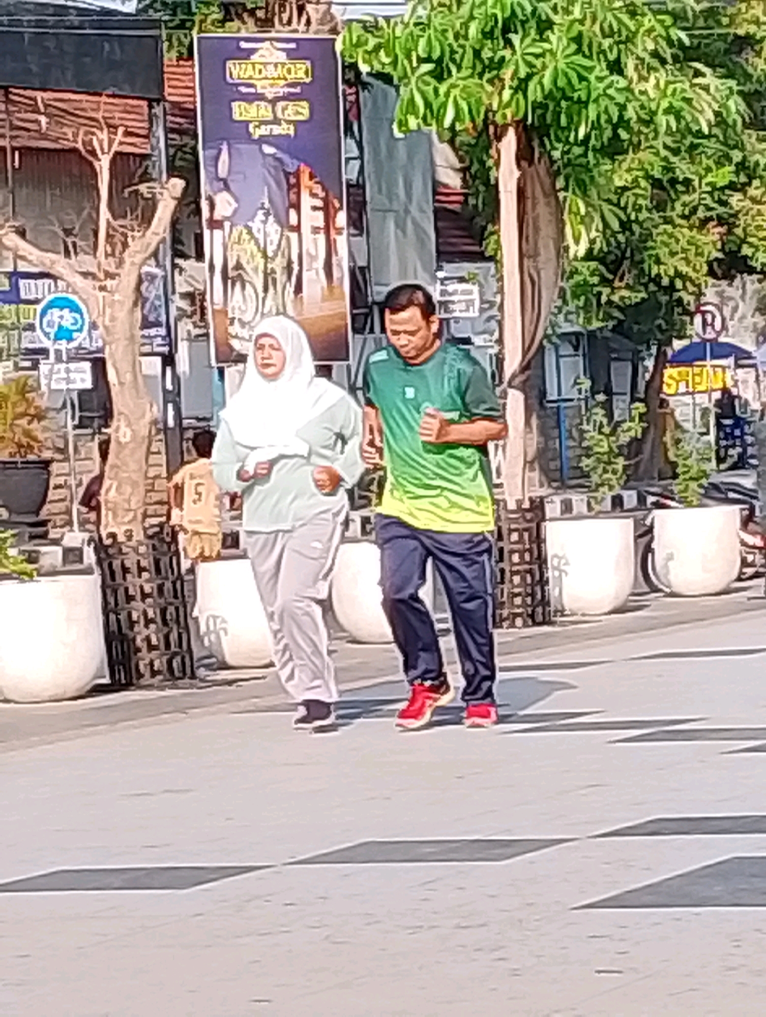 In Probolinggo, Indonesia, two individuals are captured in mid-stride during a morning jog along a paved street. The woman, on the left, wears a light green long-sleeved shirt and gray athletic pants, complemented by a white hijab. The man, on the right, sports a green and yellow gradient t-shirt with dark blue athletic pants and red athletic shoes. Both are in motion, suggesting an active lifestyle. Behind them, a banner with Indonesian text and an image of traditional Indonesian attire hangs vertically from a pole. The environment includes trees with lush green foliage, large white planters, and what appears to be a cityscape in the background, indicating an urban or semi-urban setting. The pavement is marked with dark gray lines, possibly delineating pedestrian crossings or traffic lanes. The lighting suggests it is daytime, with the sun casting shadows.