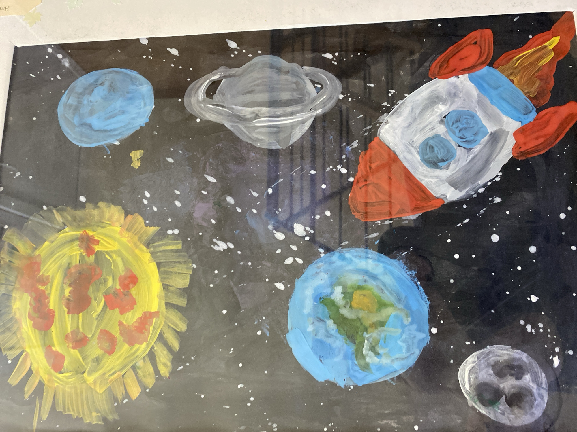 This is a photograph of a painting depicting a scene in outer space. The painting is rendered in bright, bold colors, with visible brushstrokes, suggesting it was created by a child. The primary elements are a yellow and orange sun with fiery rays, a blue and green Earth, a white and grey moon with dark craters, a red, white, and blue rocket ship with orange flames emanating from its engines, a silver flying saucer, and a blue planet. The background is black, speckled with white dots representing stars. The overall impression is one of a whimsical and imaginative portrayal of space exploration. There is no indication of location in Kaliningrad, Russia.