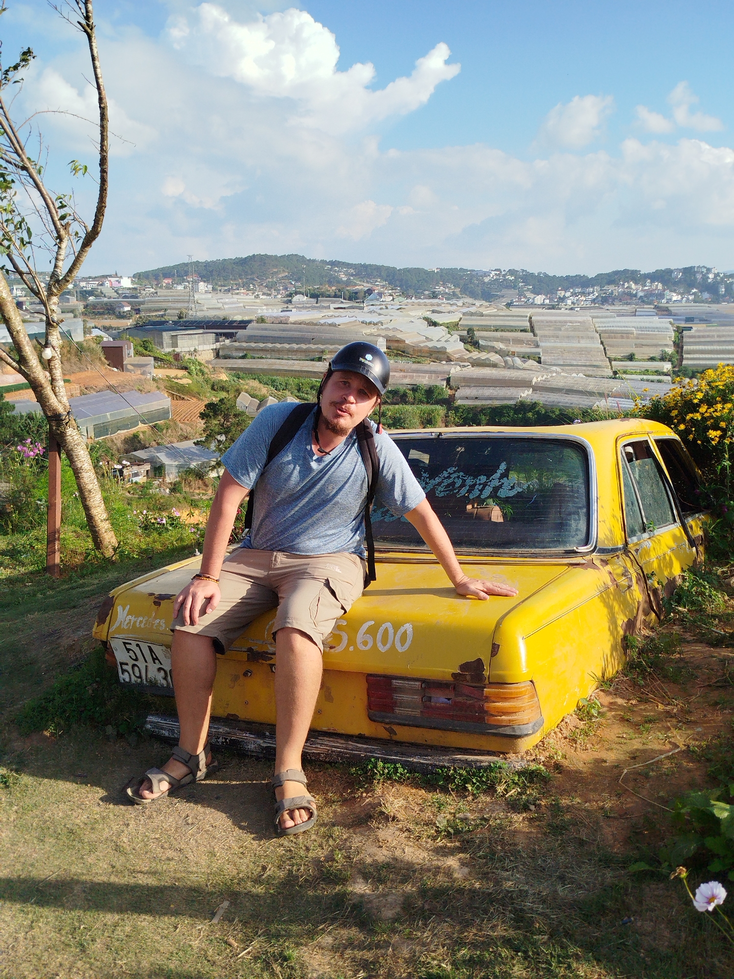 A Caucasian man with light brown hair and a goatee is seated on the trunk of a weathered, yellow Mercedes-Benz sedan. He is wearing a blue t-shirt, khaki shorts, sandals, and a black helmet. He has a backpack on his shoulders and is looking at the camera with his lips puckered. The car is positioned in an outdoor setting with a sprawling landscape of greenhouses and buildings in the background, characteristic of the Lâm Viên area in Đà Lạt, Vietnam. The foreground consists of grass and dirt, with a tree to the left and yellow flowers to the right. The sky is blue with scattered white clouds. The license plate of the car is partially visible, showing characters "51A 59436" and "5.600". The car shows signs of age and disrepair, with rust and peeling paint. The overall scene suggests a tourist attraction or a unique photo opportunity in the hilly terrain of Đà Lạt.