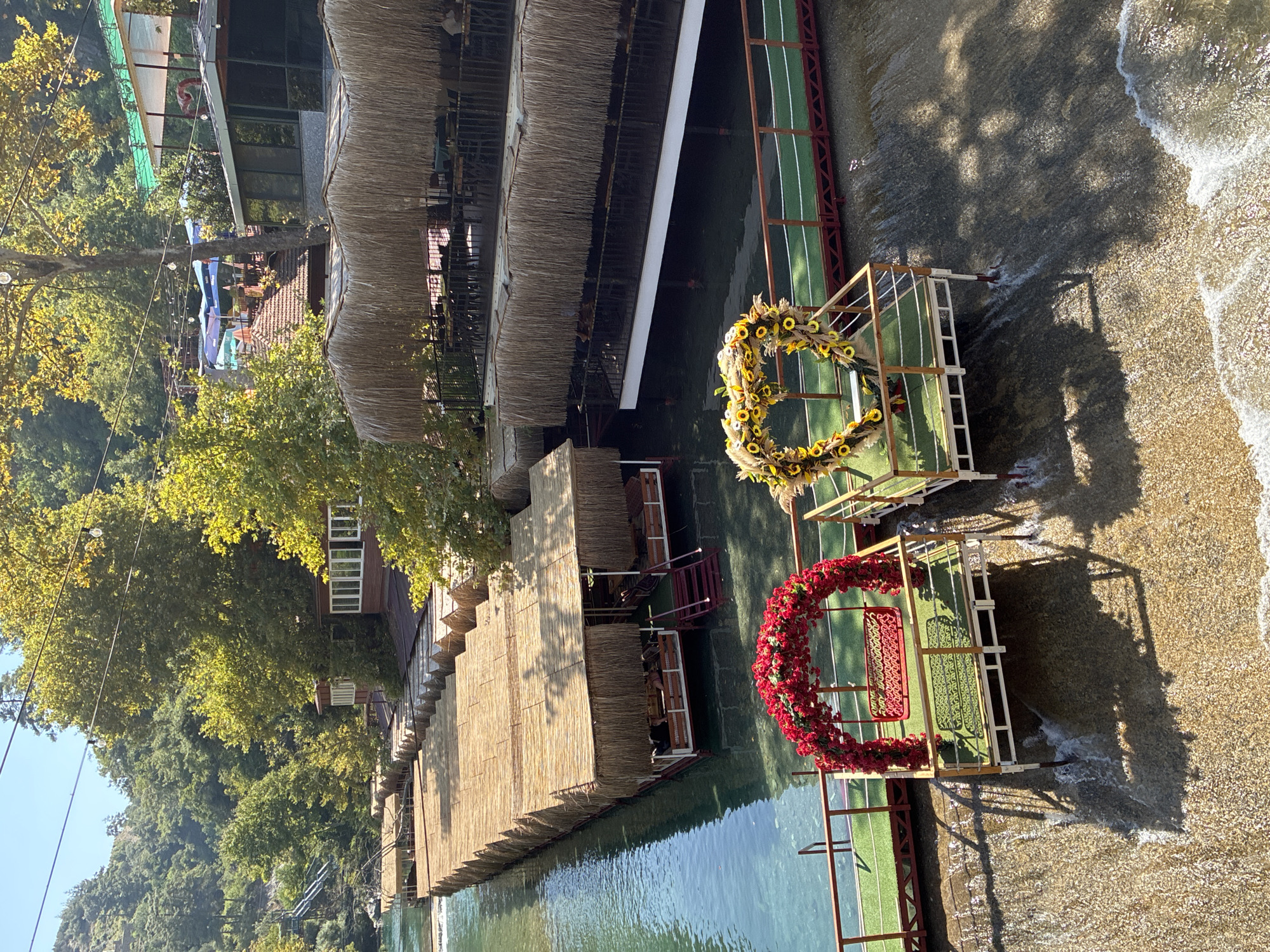 The image depicts a scenic outdoor seating area alongside a clear, shallow river, likely a part of the Dim River in Alanya, Turkey, known for its riverside restaurants. The foreground features two decorative platforms on a wooden walkway over the water. The left platform has a swing decorated with a red floral arch, and the right platform is adorned with a heart-shaped floral arrangement of yellow sunflowers. The walkway leads to a series of covered dining areas built over the water. These structures have thatched roofs and wooden supports, with tables and chairs visible within. Beyond the dining areas, the river flows downstream, with lush green trees and hills forming the background. The sunlight casts dappled shadows on the water and walkway, suggesting it is a bright, sunny day. No people are clearly identifiable in the image.