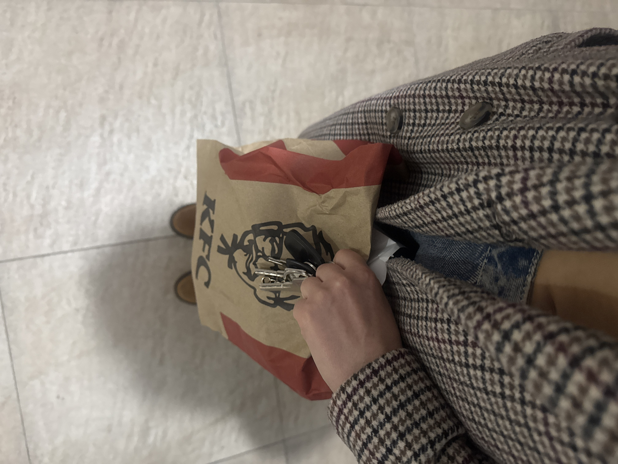 A high-angle shot shows an individual holding a KFC paper bag while standing on a tiled floor. The person is wearing a brown and black checkered coat over denim shorts. One hand is visible, reaching into the KFC bag, holding a set of keys. The tiled floor is light-colored. The location is unknown.