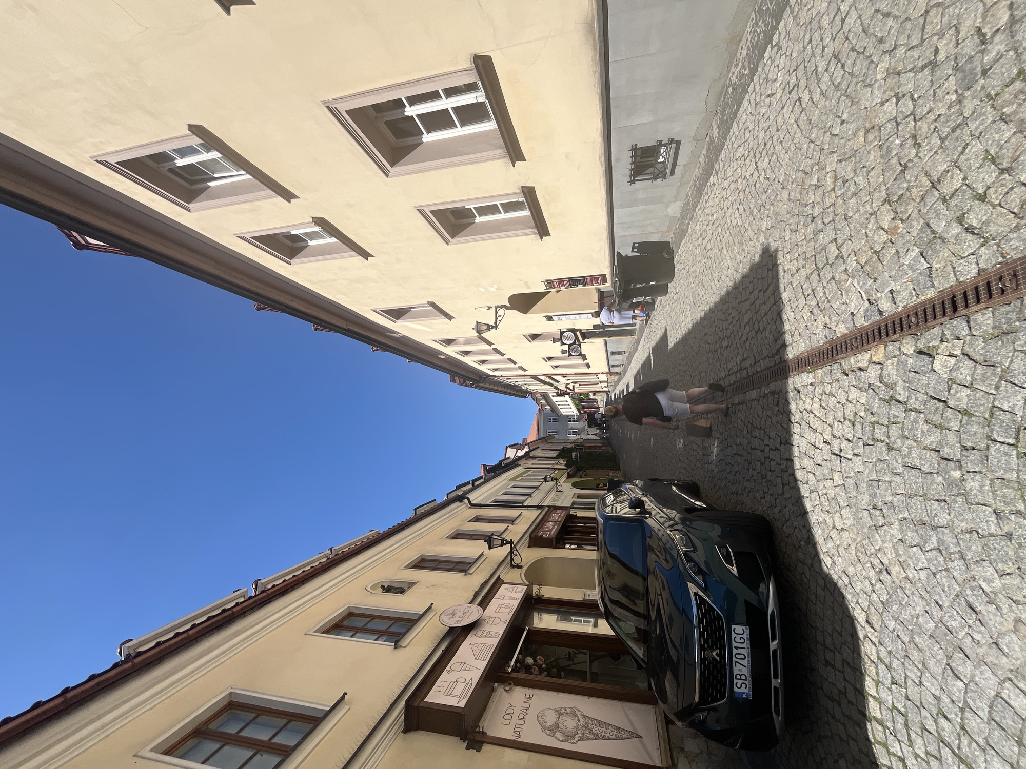 A downward-angled shot shows a cobblestone street lined with cream-colored buildings. The buildings have several windows, some with decorative trim. A dark-colored car is parked on the right side of the street. A person is walking away from the camera on the sidewalk. The sky is a clear blue. The lighting suggests it is daytime, likely sunny. Visible text includes "LODY NATURALNE" and "BISCUTIA" on signage for shops. The license plate on the car reads "SB 701GC". The scene appears to be a historic European street.