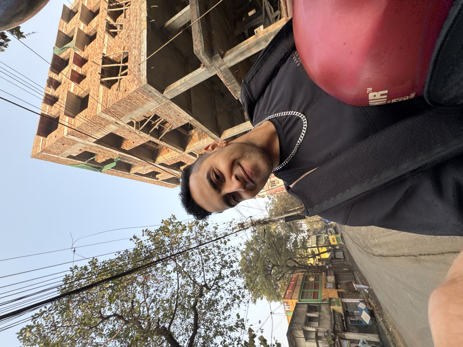 A man smiles at the camera while presumably riding a motorbike, wearing a black shirt and silver chain. He has a helmet on. The background includes an unfinished brick building, trees with bare branches, utility lines, and a street with other buildings. The photo is taken from an unusual angle, tilting the scene sideways.