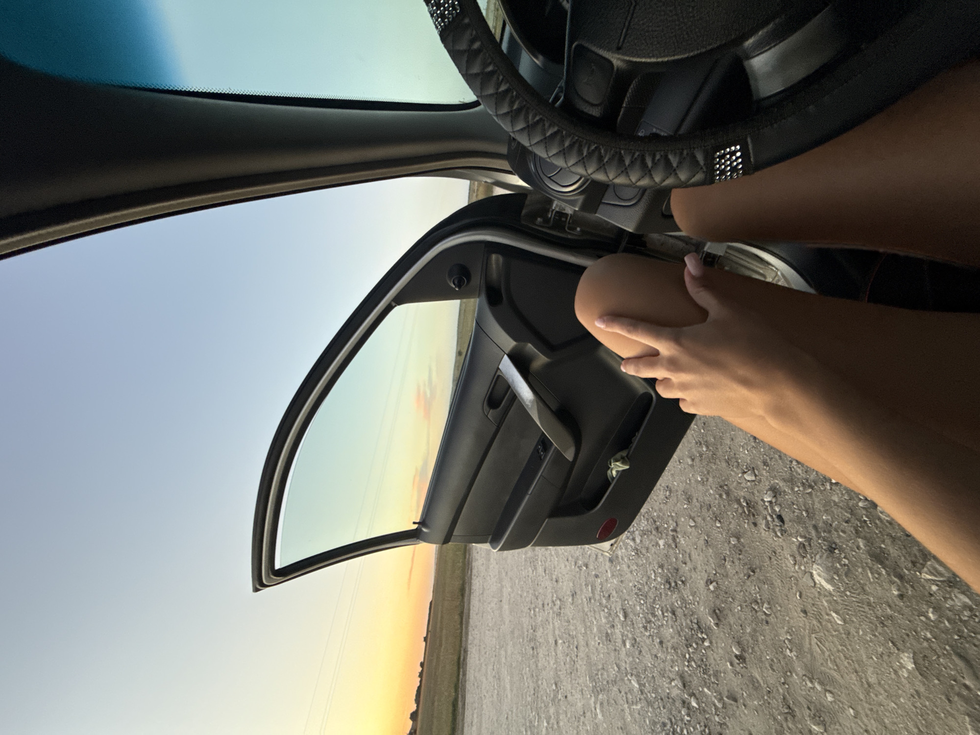 The image shows a person's legs resting against the open car door of a vehicle parked on a gravel road. The sun is setting in the background, casting an orange glow over a field. The interior of the car is visible, including the steering wheel and the car door panel. The scene appears to be a rural area, possibly outside of Cimișlia, Moldova, given the natural landscape.