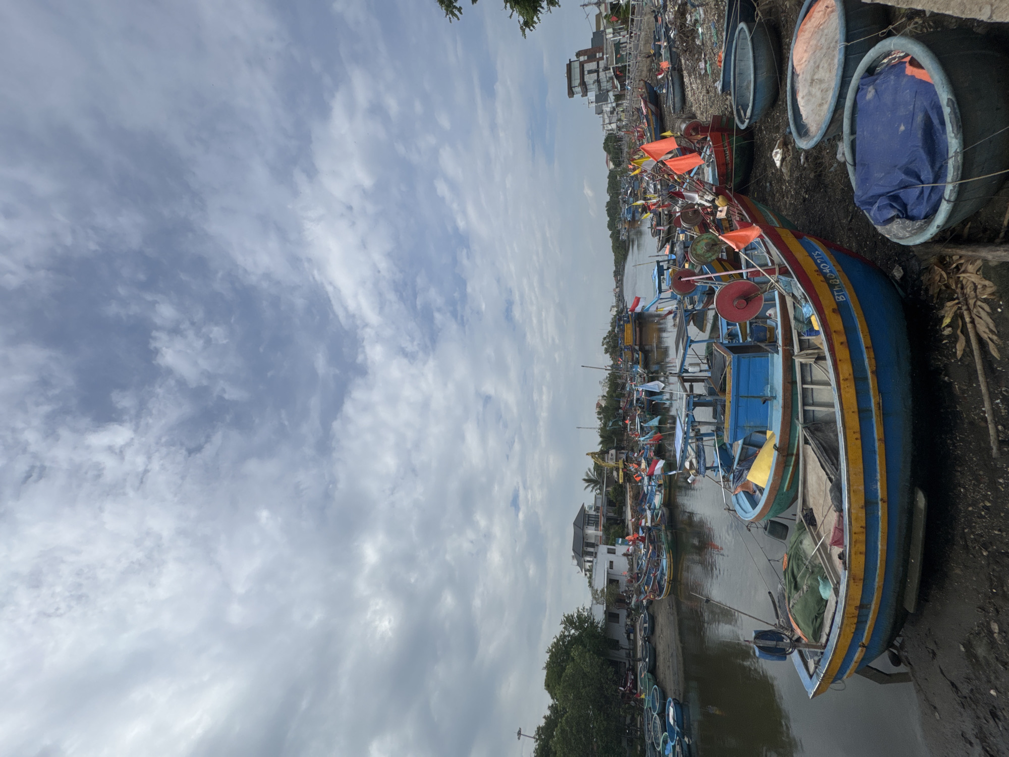 The image depicts a waterfront scene in Phú Thủy, Vietnam, characterized by a multitude of fishing boats docked along a riverbank. The boats, primarily small to medium-sized, are brightly colored with yellow and blue being prominent. Many are adorned with flags, some red and white, others orange, suggesting a maritime tradition. Nets and fishing gear are visible on and around the vessels. The river itself appears calm, reflecting the cloudy sky above. On the riverbank, there are signs of human habitation and activity, including houses and what appears to be a construction site with a yellow excavator in the distance. The foreground shows the muddy bank of the river with several round, basket-like containers, some covered with tarps or containing fishing nets. The overall atmosphere is one of a working fishing village, with a mix of natural elements and human infrastructure under a predominantly cloudy sky.