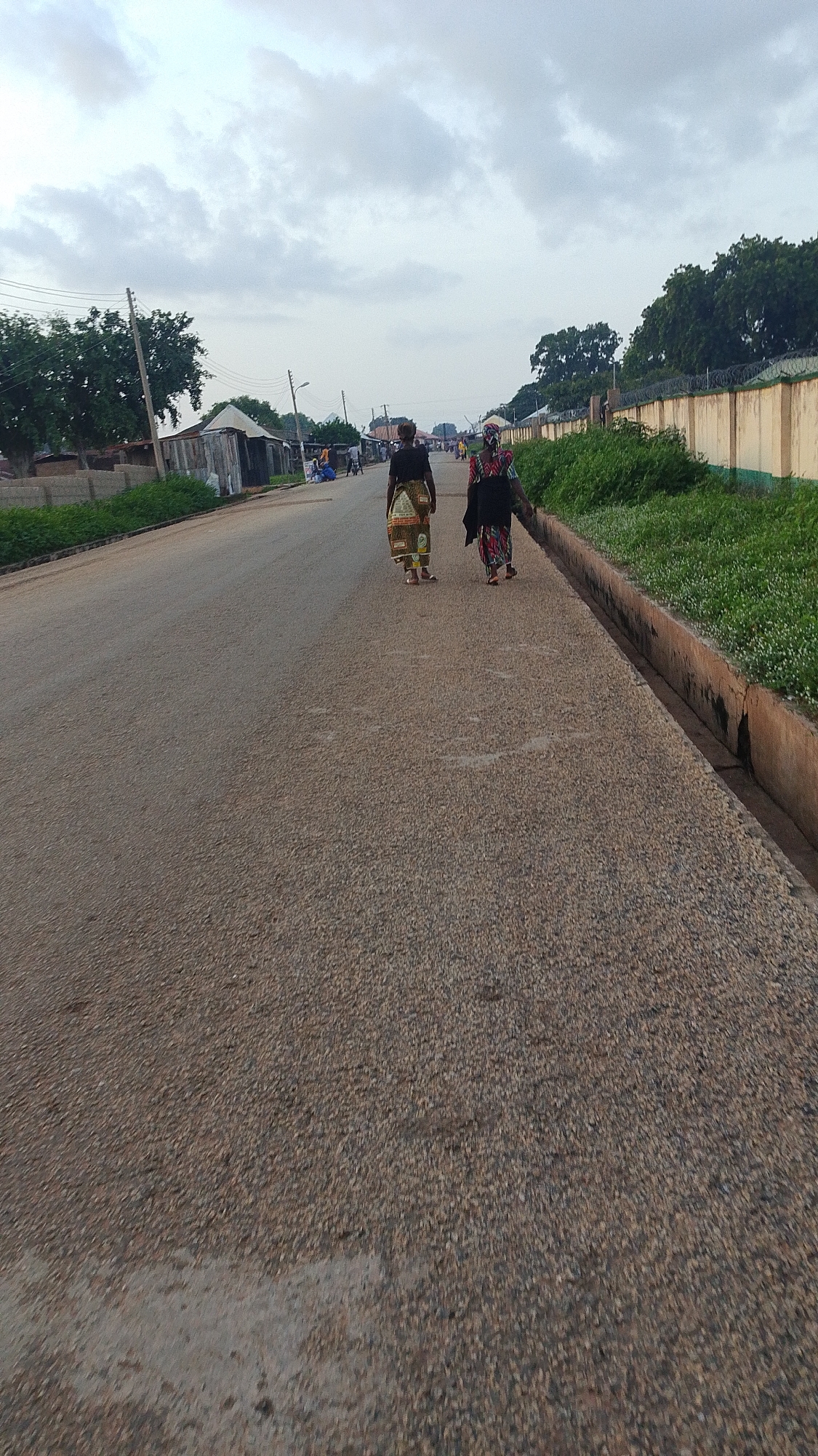 The image depicts a street scene in Donga, Nigeria, under an overcast sky. Two women are walking away from the camera down a paved road. The woman on the left wears a patterned skirt and dark top, while the woman on the right wears a multi-colored patterned dress and a headscarf. To the right of the road, a concrete drainage channel runs alongside a grassy area bordered by a yellow wall with barbed wire on top. On the left side of the road, several structures are visible, including what appear to be small buildings or stalls and some trees. The road is made of asphalt with visible aggregate. In the background, other people and structures are faintly discernible, suggesting a populated area.