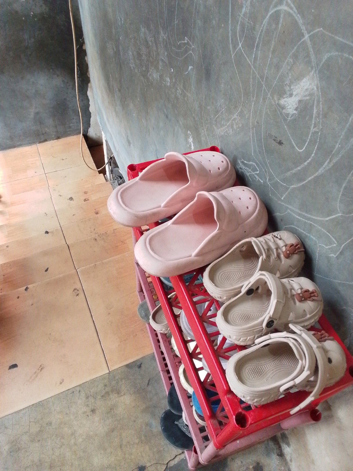 A pair of pale pink slip-on shoes and two pairs of light beige clogs are displayed on a red plastic shoe rack. The shoe rack is positioned against a textured, grey concrete wall with white chalk scribbles. To the left of the shoe rack is a floor covered with light brown tiles. A beige electrical cord runs vertically down the wall, starting from the top left corner of the image. The scene appears to be indoors, likely in a residential setting.