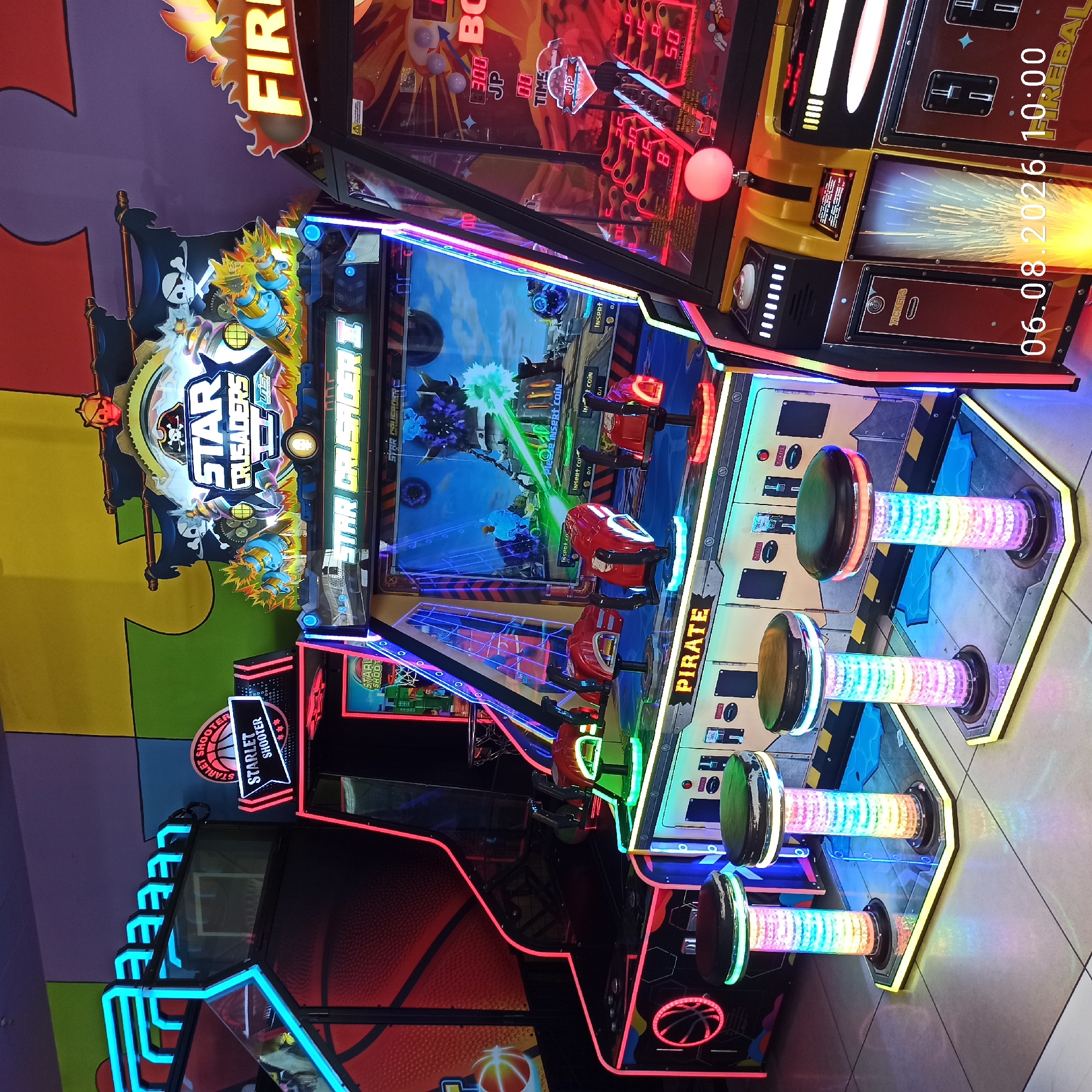 The image depicts an indoor arcade in Bari, Italy, featuring several illuminated video game machines. In the foreground, three colorful, cylindrical stools with black seats are visible, each illuminated by internal rainbow LED lights. Behind the stools are two arcade machines. The machine on the left is a "Starlet Shooter" basketball game with neon blue and red lighting. To its right is a "Star Crusader II" arcade game, characterized by a large screen displaying gameplay with pirate and space-themed graphics, and two mounted red gun controllers. Further to the right, a portion of a "Fireball" arcade machine is visible, with prominent yellow and red lighting. The background consists of a wall with large, brightly colored rectangular panels in yellow, green, purple, and blue. The floor is tiled. A timestamp "06.08.2026 10:00" is superimposed on the lower right corner of the image. There are no people visible in the scene.