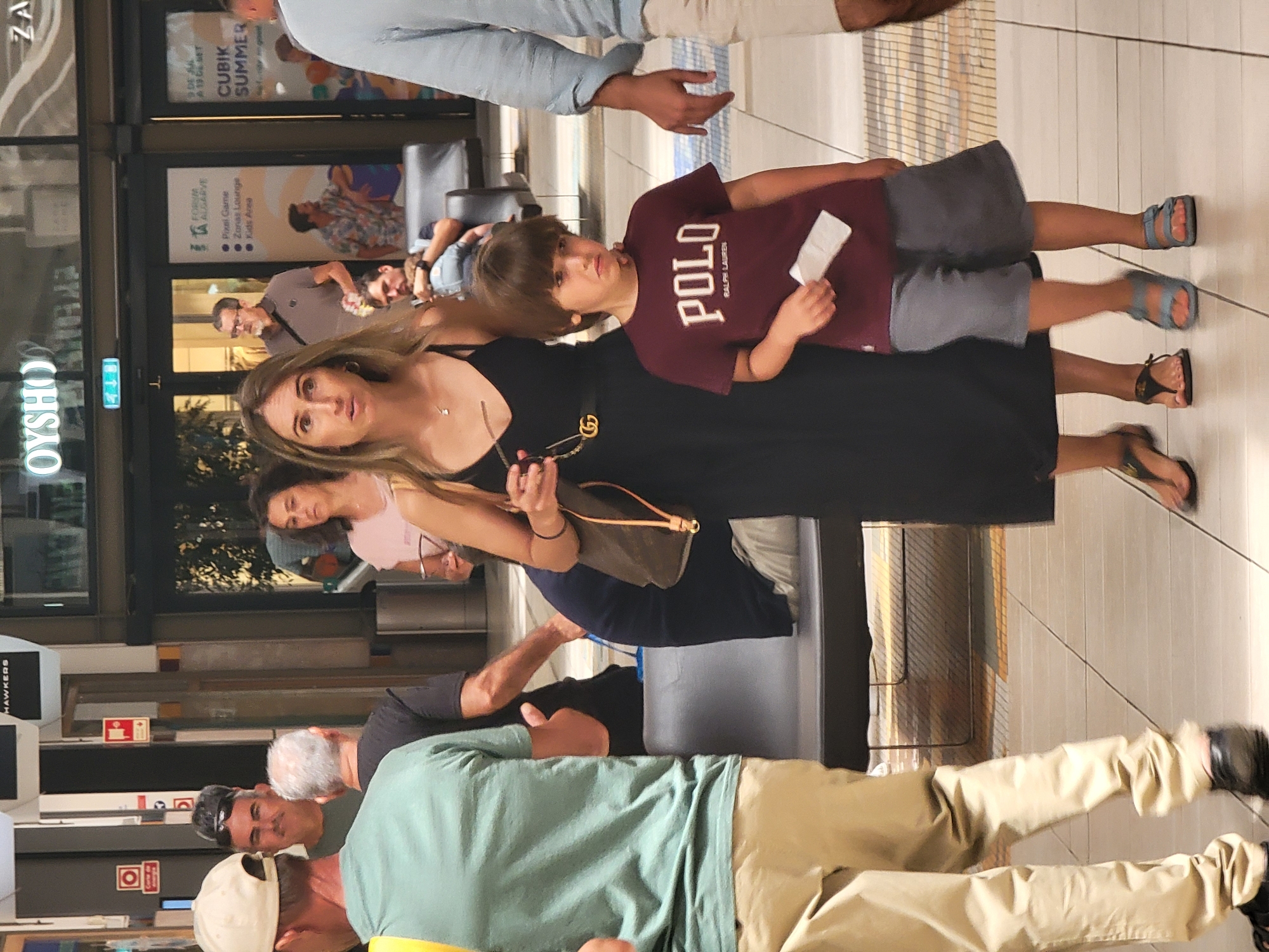 This is a full-body, slightly low-angle shot of a group of people in an indoor public space that appears to be a shopping mall. In the foreground, a young boy wearing a maroon Polo Ralph Lauren t-shirt and grey shorts stands looking towards the camera with a neutral expression. He is holding a white slip of paper. To his left is a woman in a black dress and to her left is a man in a light green t-shirt and khaki pants. Behind them, more people are visible, some in the background out of focus. The environment includes tiled flooring and shops with signage, including "OYSHO" and "CUBIK SUMMER". The overall impression is a candid snapshot of individuals navigating a busy commercial area. Based on the signage "FORUM ALGARVE" visible in the background, this location is identified as Forum Algarve, a shopping mall in Faro, Portugal.
