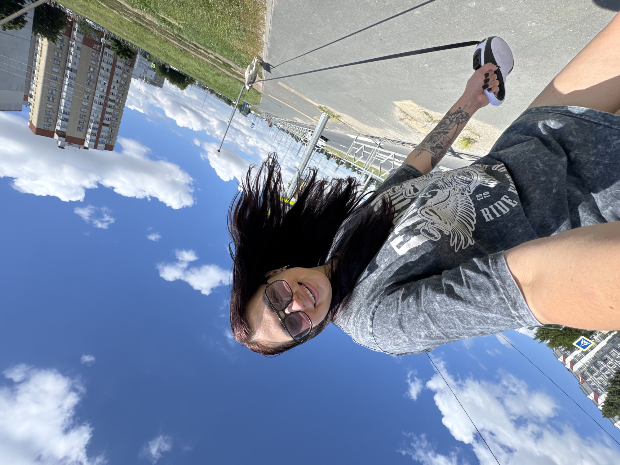 A woman with long dark hair and sunglasses smiles at the camera, holding a retractable dog leash. She wears a gray t-shirt dress with a graphic print. Her right arm has a tattoo. She is standing on a paved path, possibly a sidewalk or bike lane, in a city. To her right, a light-colored dog walks on the leash. In the background, there is a tall apartment building with red and white trim, a grassy area, a road with vehicles, and a bright blue sky with white clouds. The image appears to have been taken outdoors in Tolyatti, Russia.