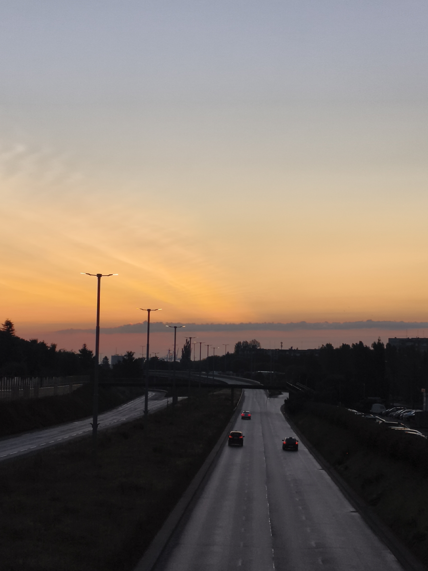A vertical, medium-range shot captures a highway scene at dusk in Gdansk, Poland. The sky transitions from a pale blue at the top to a vibrant orange near the horizon, with faint clouds. Tall streetlights with bright, white bulbs line the left side of the highway, their light reflecting on the wet asphalt. The highway curves to the right, with three cars visible: a red one in the distance, a yellow one in the middle lane, and a black one in the right lane. Trees and vegetation form dark silhouettes on the right and left sides of the highway, with buildings and a power line visible in the distance on the right. A parking lot filled with cars is also visible on the far right.