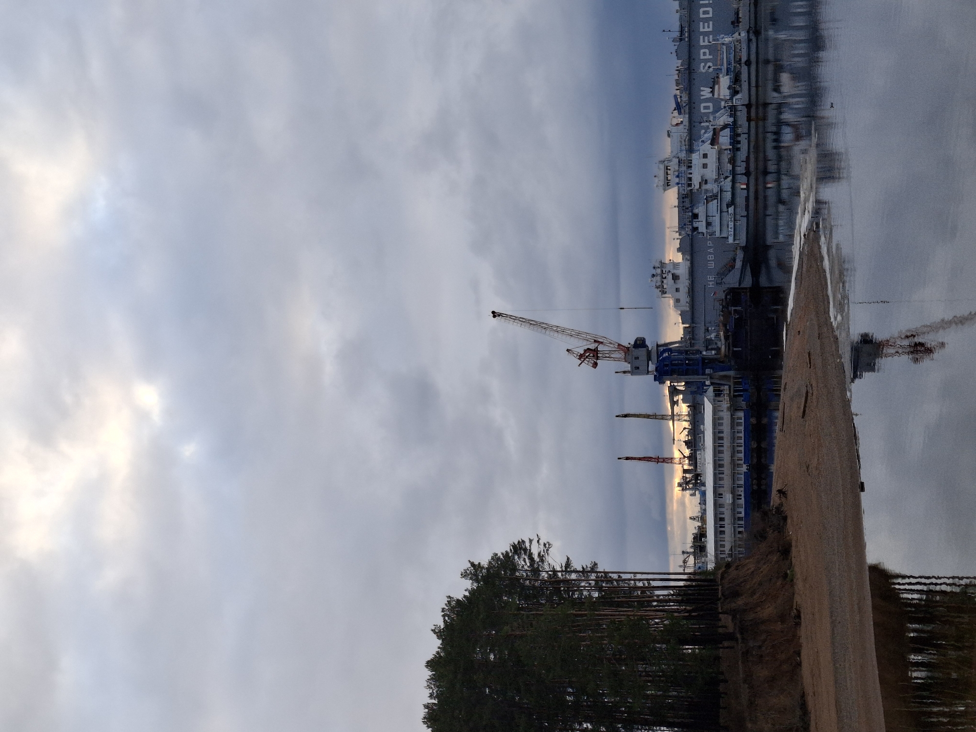 The image depicts a waterfront scene in Chaikovsky, Russia, under an overcast sky, with soft light indicating late afternoon or early evening. The view spans across a body of water to an industrial area on the opposite bank.
In the foreground, a dark stand of coniferous trees anchors the left side, with a sandy or earthy bank sloping gently to the water's edge.
Across the calm water, a port or industrial facility is visible. Several large ships are docked along the far shore. A prominent ship clearly displays "SLOW SPEED!" in white lettering, with "НЕ ШВАРТОВАТЬ" (Do Not MoOR) and the number "374 085" also visible below it. Various buildings, likely part of a shipyard or factory, line the distant shore.
Multiple cranes punctuate the skyline of the industrial area; one notable red and white crane is positioned towards the center. The sky is predominantly covered by light grey clouds, though patches of brighter, diffused light are visible on the left. The water's surface provides a mirror-like reflection of the sky and the distant structures, contributing to the tranquil yet industrial atmosphere.