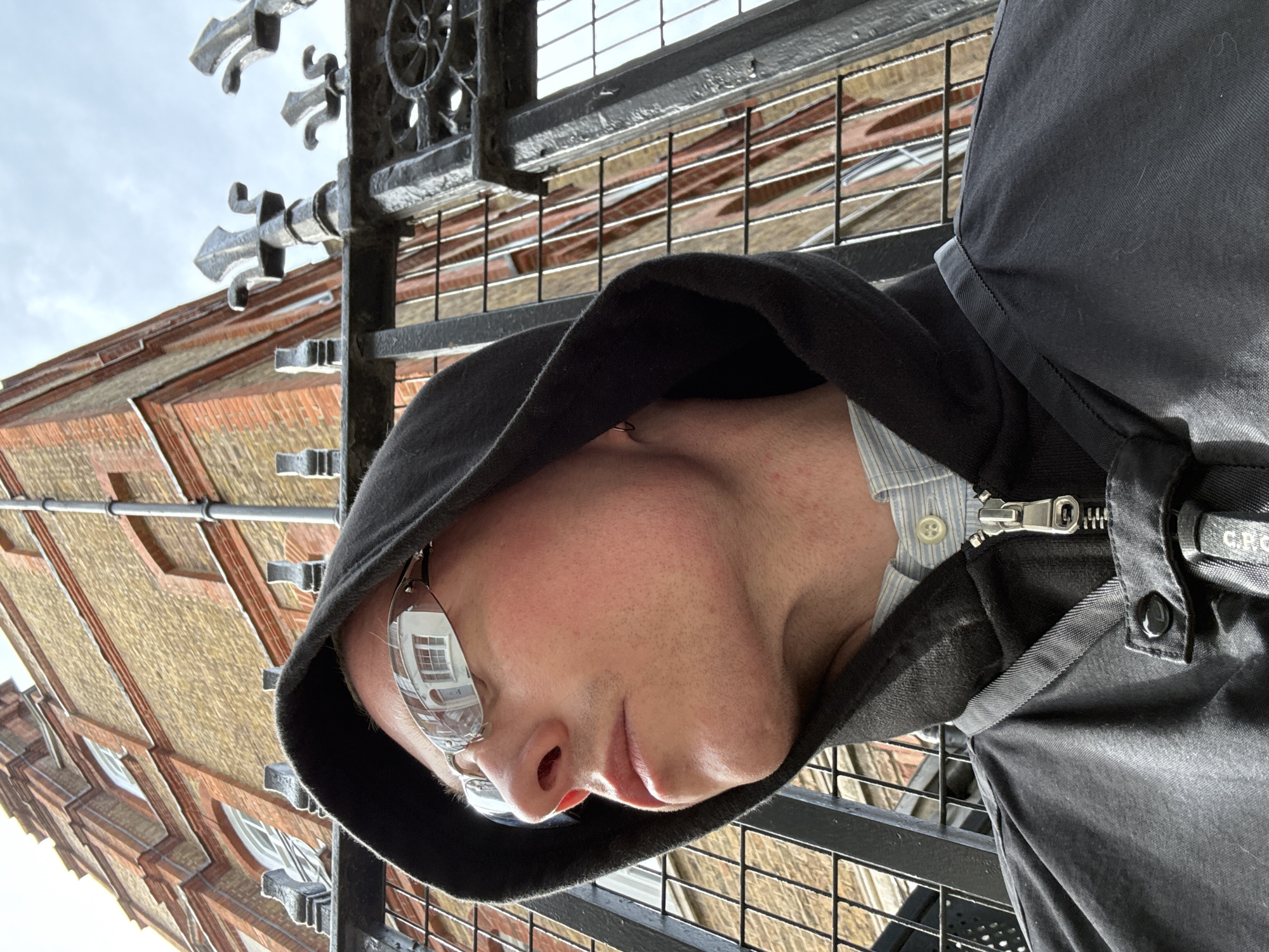 A medium, close-up, rotated-counterclockwise, outdoor shot of a person wearing a hooded jacket and glasses standing in front of a building and gate in what appears to be London, United Kingdom. The person is wearing a dark-colored hooded jacket over a light-colored, button-down shirt. The jacket is zipped, and a detail on the jacket near the zipper is visible. The person is also wearing glasses with light-colored frames. The reflection in the glasses shows a window and what appears to be the interior of a building.
The person is standing in front of a dark-colored metal gate. Behind the gate is a building made of light-colored bricks. The building has a pattern of darker-colored bricks around the windows. The sky is visible in the upper portion of the frame. The gate has decorative elements.