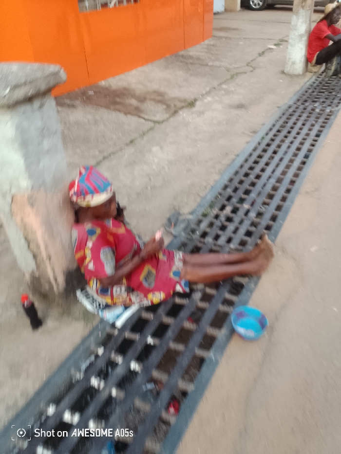 A woman is lying on a metal grate next to a street curb. She wears a red dress with a blue and pink headwrap. There is a brown cushion behind her. A Coca-Cola bottle is to her left. A small blue bowl is to her right, near her feet. In the background, a man in a red shirt is seated. An orange building and a parked vehicle are also visible. The words "Shot on AWESOME A05s" are at the bottom of the image.