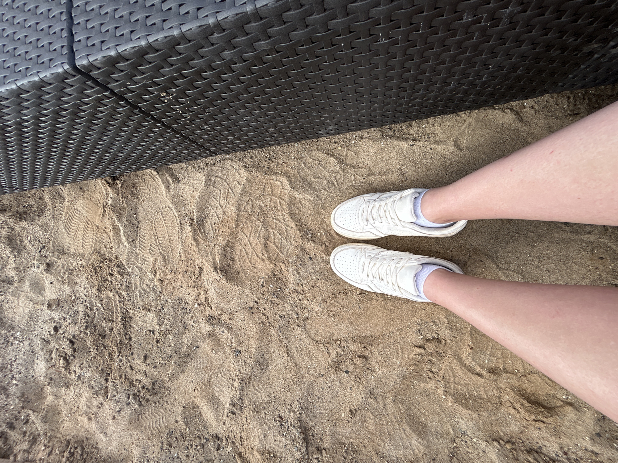 The image shows a person's legs and feet wearing white sneakers and white socks, standing on a sandy surface. To the right of the sand is a dark, woven textured surface, possibly a piece of furniture or outdoor seating. The sandy area shows tire tracks or shoe imprints. The lighting suggests it is daytime. The scene is not definitively located in Chaikovsky, Russia.