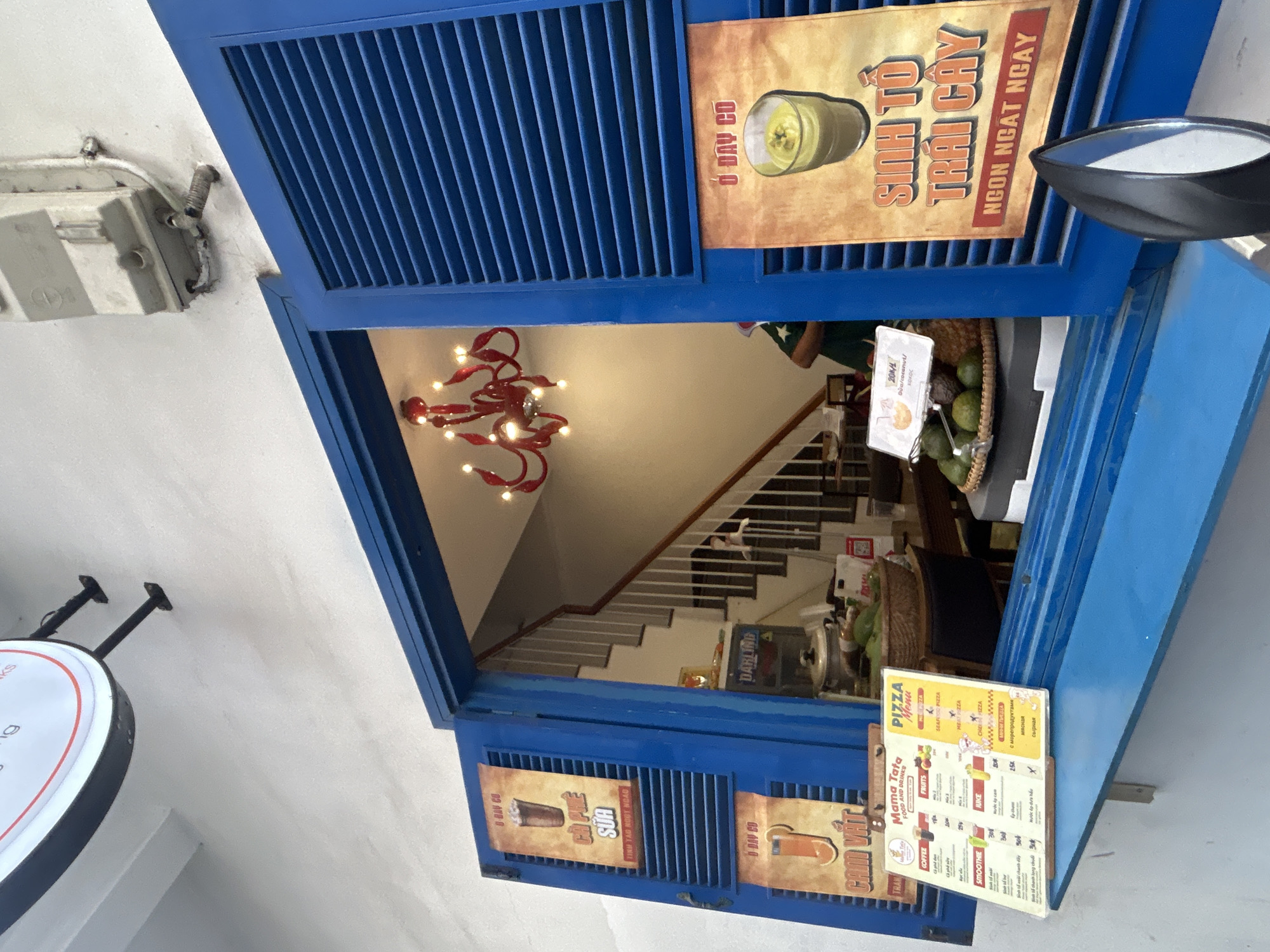 A shot taken from outside a business shows the open blue shutters of the shop's front, revealing an interior with a staircase. The interior is lit by a chandelier. A menu is displayed on the shutter, advertising pizza, coffee, smoothies, and fruit drinks. Signs in Vietnamese advertise Sinh To Trai Cay and other drinks. A sign inside the shop reads "Darling." The sign mentions Mama Tata Food and Drinks and that coconuts cost 20K. The presence of Sinh To Trai Cay suggests this is Nha Trang, Vietnam.