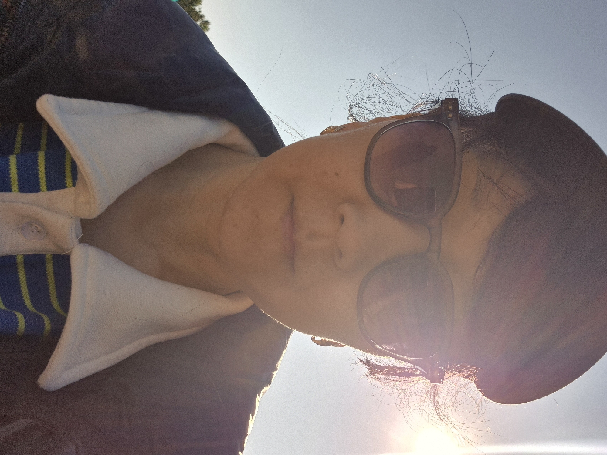 A woman with sunglasses and a hat is looking into the camera. She is wearing a shirt with a white collar and blue and yellow stripes. The background is a bright sky. The location appears to be outdoors, but no specific landmarks indicating Kokshetau, Kazakhstan, are visible.