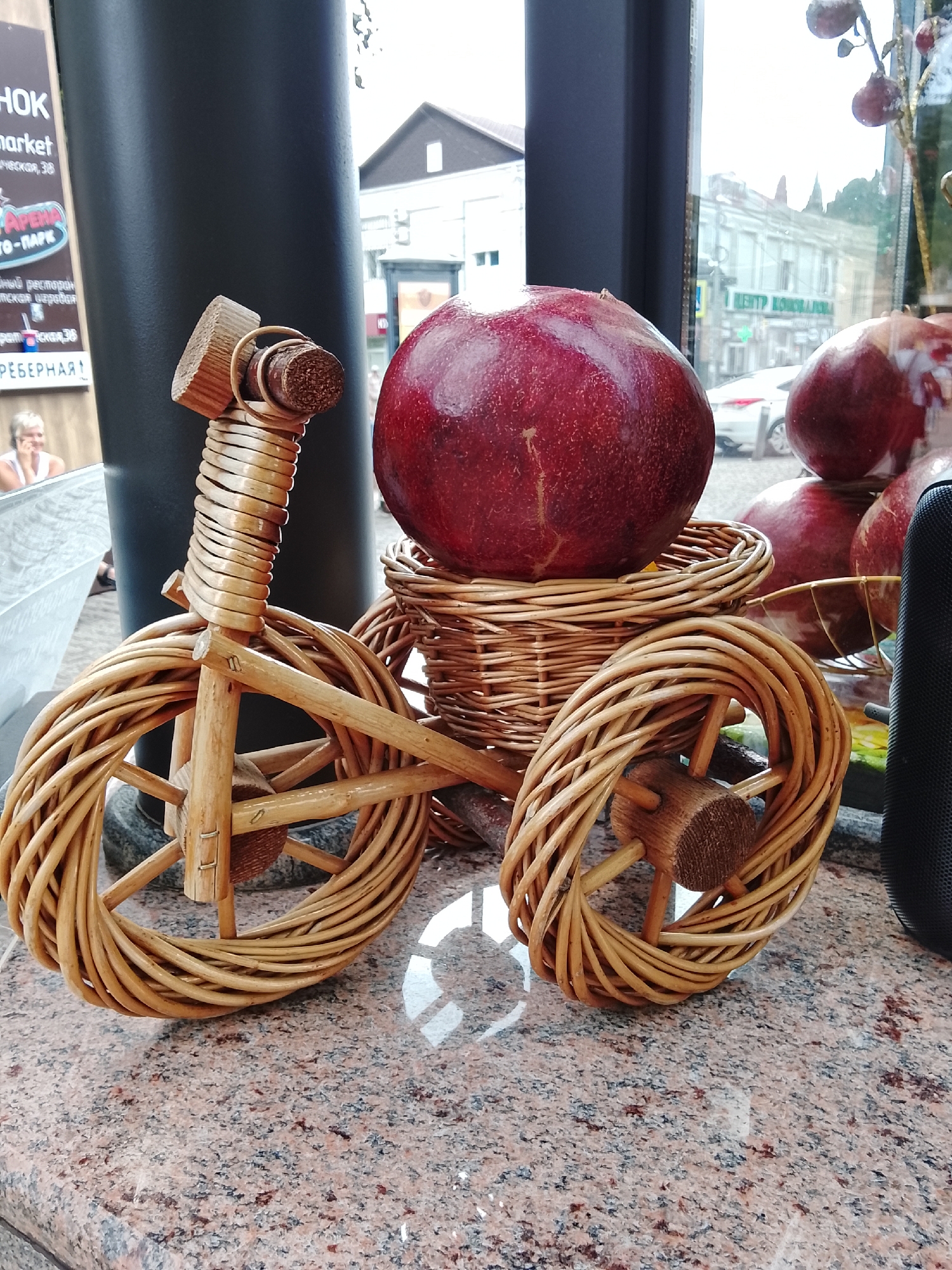 A wicker tricycle with a large red fruit, likely a pomegranate or passion fruit, placed on its basket is positioned on a granite countertop. The tricycle has a wooden frame and wicker wheels. In the background, visible through a window, is a street scene in Sochi, Russia, with buildings and a car. The sunlight reflects off the polished countertop. A person can be seen blurred in the far left of the image.