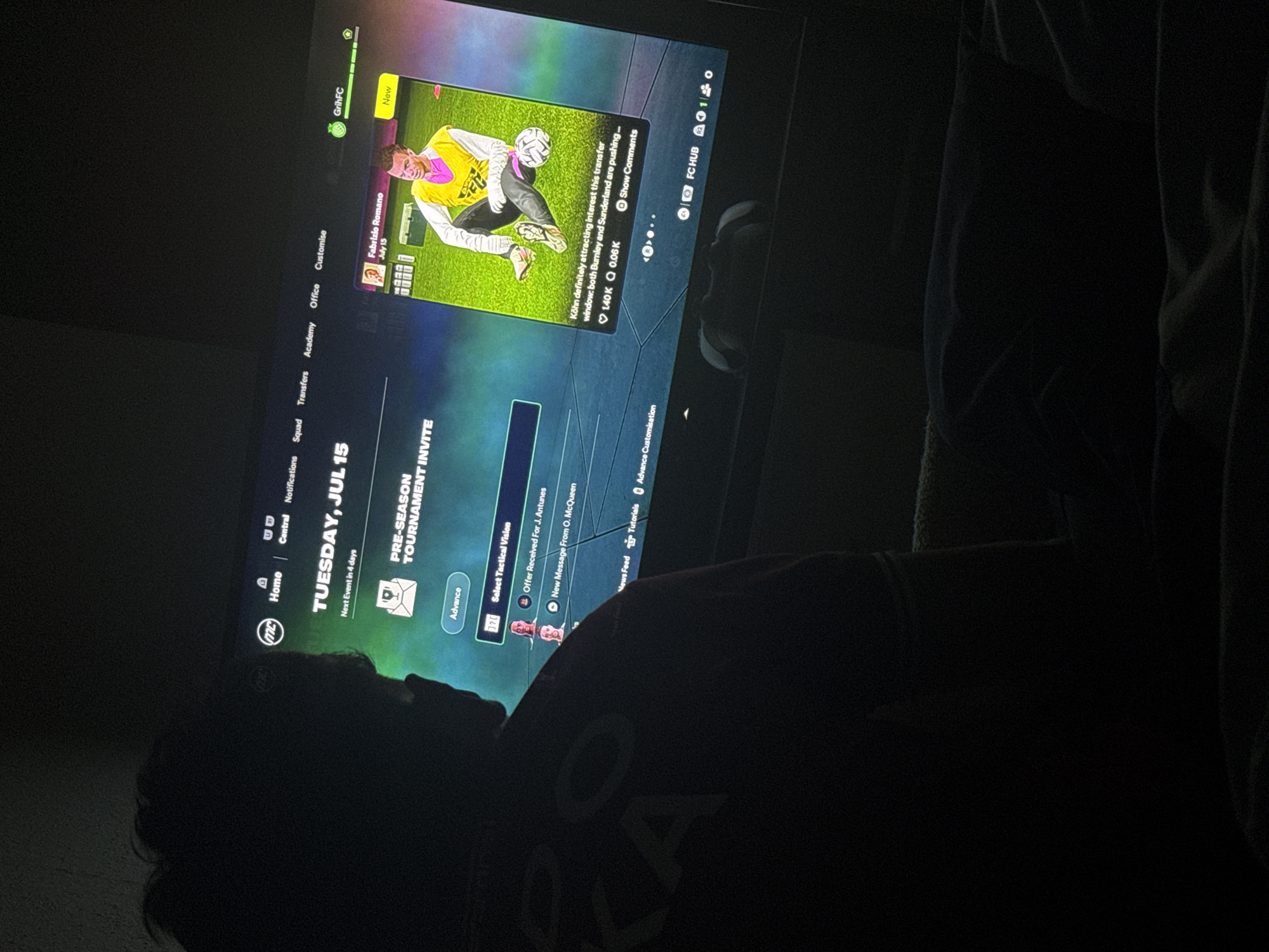 The image shows a person sitting in a dark room, likely at home, playing a soccer video game on a large screen TV. The game interface is visible, showing team management options, notifications, and a report about player interest from other teams. The date displayed in the game is Tuesday, July 15. The person appears to be casually dressed.