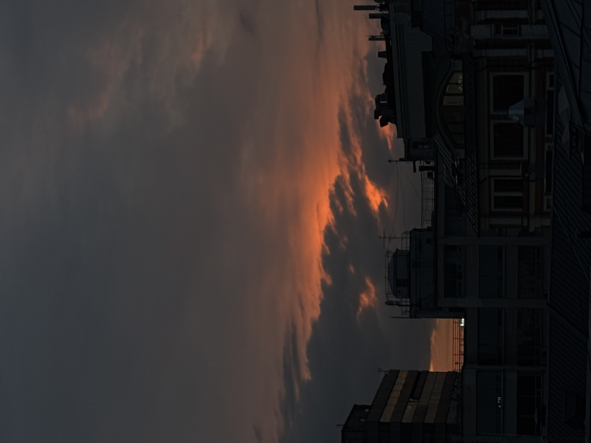 The image depicts a dark urban cityscape at dusk, likely in Moscow, Russia. The sky is dominated by heavy, dark clouds tinged with orange and red light from the setting sun. Silhouetted buildings are visible against the skyline, including what appears to be Soviet-era construction. No people are discernible. The overall impression is of a cold, dark, industrial city at twilight.