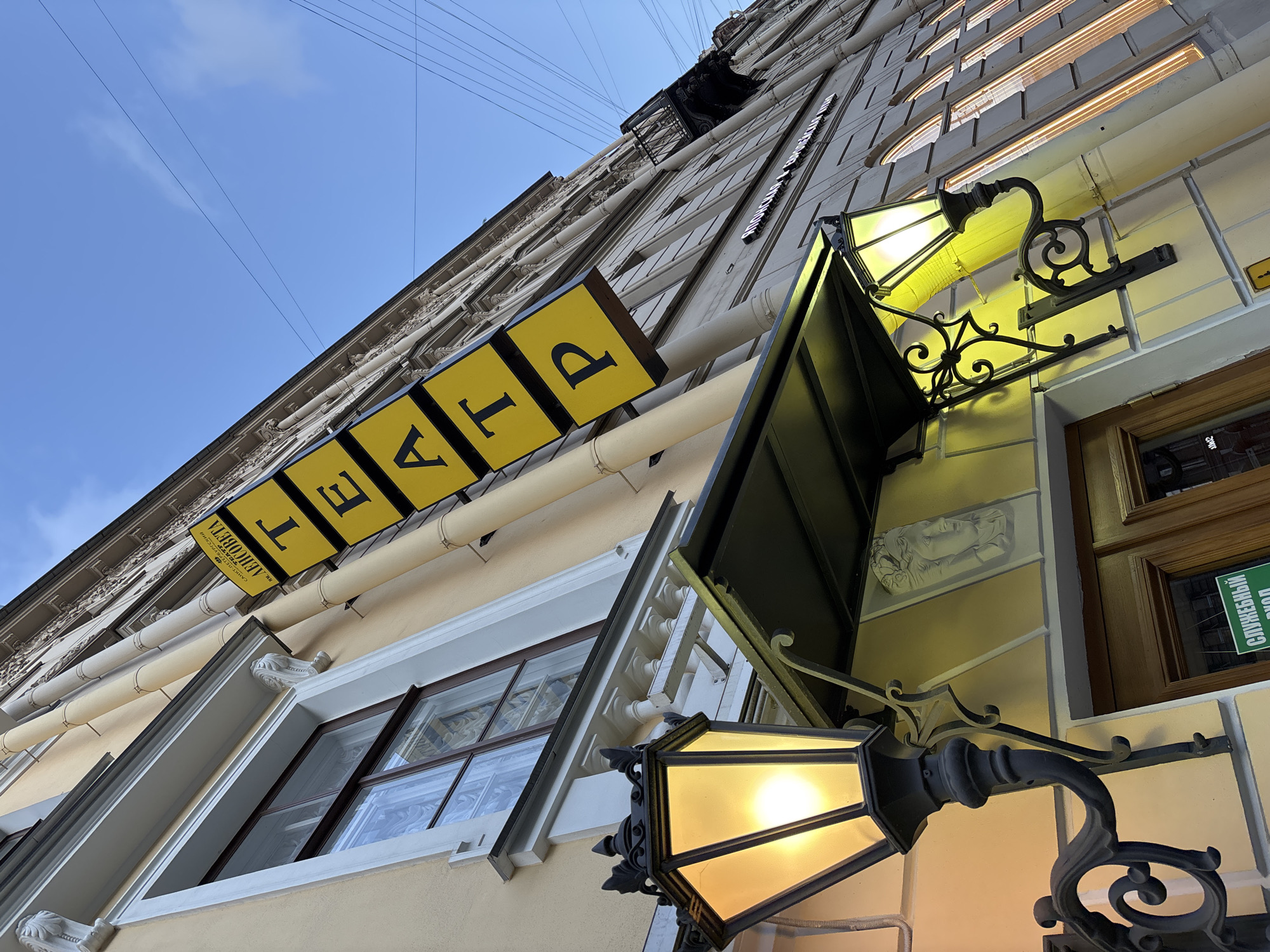 A low-angle shot captures the ornate facade of a building, likely a theatre, under a clear blue sky in Saint Petersburg, Russia. The perspective is tilted, emphasizing the height of the building and its architectural details.
The building's facade is pale yellow or beige, adorned with classical moldings, cornices, and decorative elements. Light-colored pipes run horizontally across the upper levels. Visible are multiple windows with white and darker frames, some reflecting the surroundings.
Dominating the mid-ground is a large yellow sign with individual black Cyrillic letters spelling out "Т Е А Т Р" (Teatr), meaning "Theatre." Below this, a smaller white sign reads "САНКТ-ПЕТЕРБУРГСКИЙ МОРСКОЙ ДРАМАТИЧЕСКИЙ ТЕАТР им. М.Ю. БОГУСЛАВСКОГО" (Saint Petersburg Maritime Drama Theatre named after M.Yu. Boguslavsky), along with a web address "WWW.S-PETERBURG.COM".
Two elaborate, black wrought-iron lamps with frosted or yellowish glass glow brightly, casting a warm light on the wall directly beneath them. One lamp is prominent in the foreground, with its intricate details clearly visible. Between windows, near one of the lamps, a light-colored sculptural relief of a face is embedded in the wall.
In the bottom right corner, a green sign with white Cyrillic text "СЛУЖЕБНЫЙ ВХОД" (Sluzhebnyy Vkhod), indicating a "Staff Entrance" or "Service Entrance," is partially visible. Overhead, several thin wires crisscross against the sky. The bright lamps contrast with the daytime sky, suggesting they are either always on or the photo was taken at dusk/dawn.