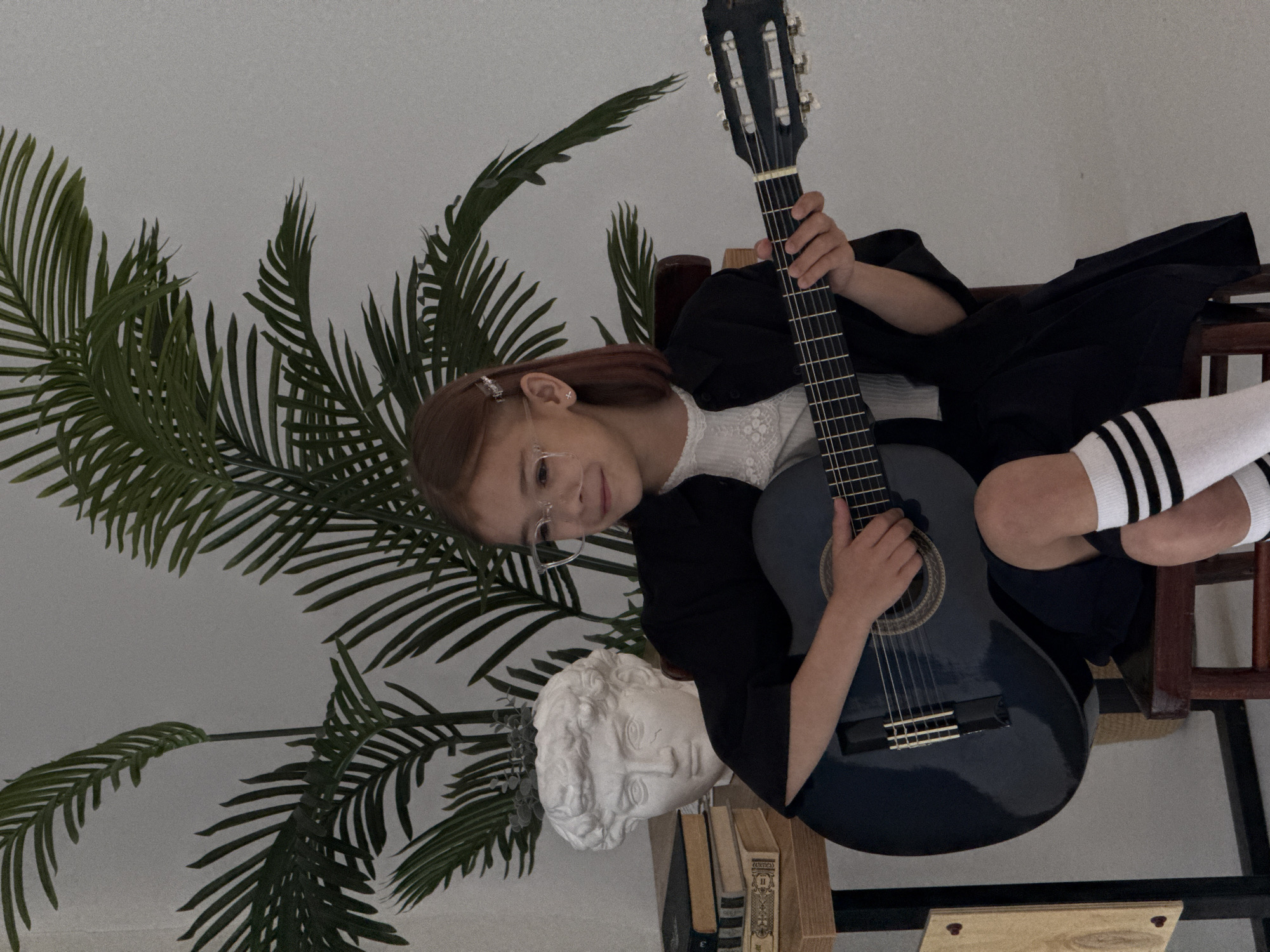 A young girl with brown hair and glasses sits indoors, holding a blue classical guitar. She wears a white top with lace details, a black jacket, and black skirt. Her legs are crossed, revealing white knee-high socks with black stripes. To her left is a potted plant with large, palm-like leaves, and a white bust resting on a stack of books. The girl is positioned on a wooden stool. There is a wooden shelf structure visible in the lower right of the image. The background is a plain white wall. The location is the city of Chaikovsky, Russia.