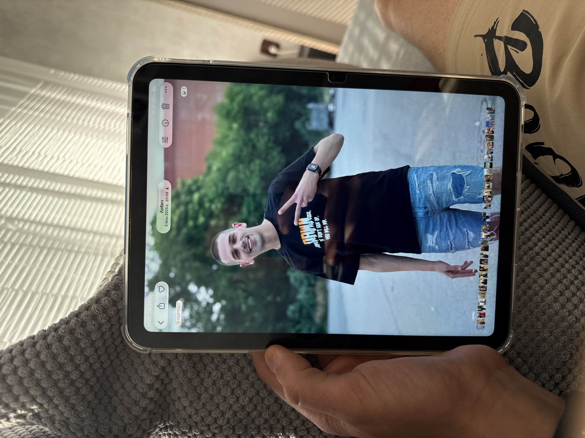 A person is holding a tablet displaying a photograph of a man outdoors. The man in the photo is making a peace sign with his left hand, wearing a black T-shirt with text, distressed blue jeans, and a watch on his left wrist. The background appears to be an urban environment with greenery. The tablet's screen shows photo editing interface elements, and the person holding the tablet is sitting on what appears to be a grey textured blanket. The text shown on the tablet is in Bulgarian and says "Lovech," a city in Bulgaria, and includes date and time information.