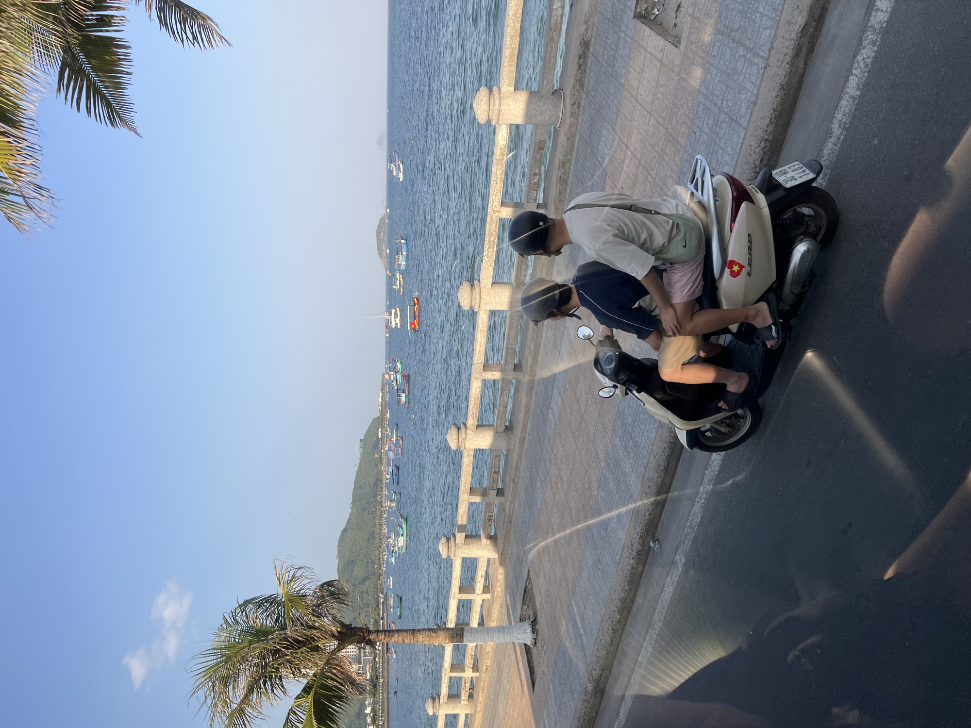 Two individuals, both wearing helmets, are riding on a beige scooter on a road alongside a promenade in Nha Trang, Vietnam. The person in front is wearing a dark blue t-shirt and the person behind is wearing a light-colored t-shirt and pink shorts. The scooter is moving away from the camera. The promenade has a stone railing and a tiled walkway. Beyond the promenade is the Nha Trang coastline, with numerous small boats in the water. In the background, there are hills and buildings. Palm trees are visible on the left side of the image. The sky is clear and blue.